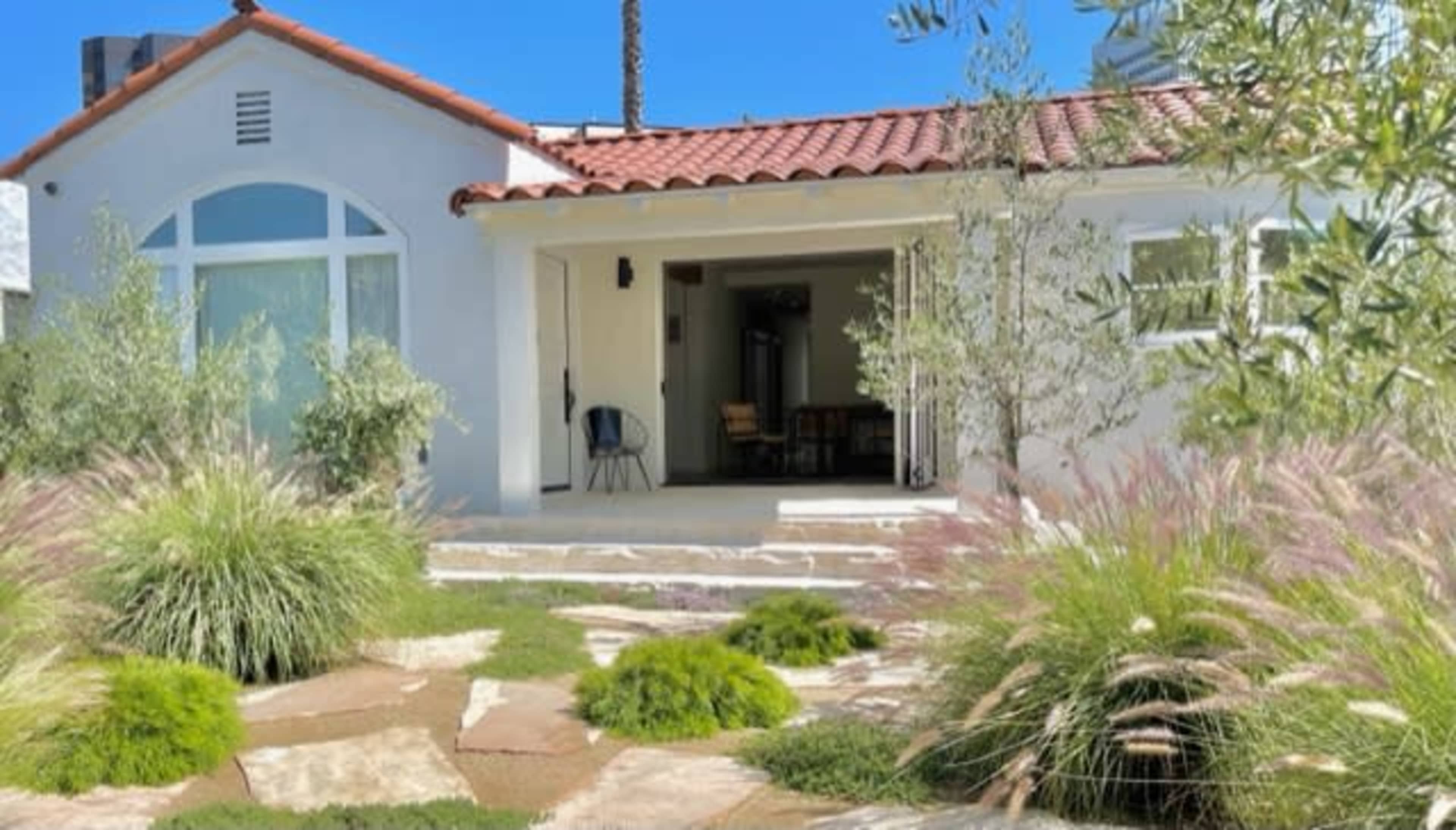 A white house with a red tile roof features a stone pathway and landscaped greenery in the front yard.