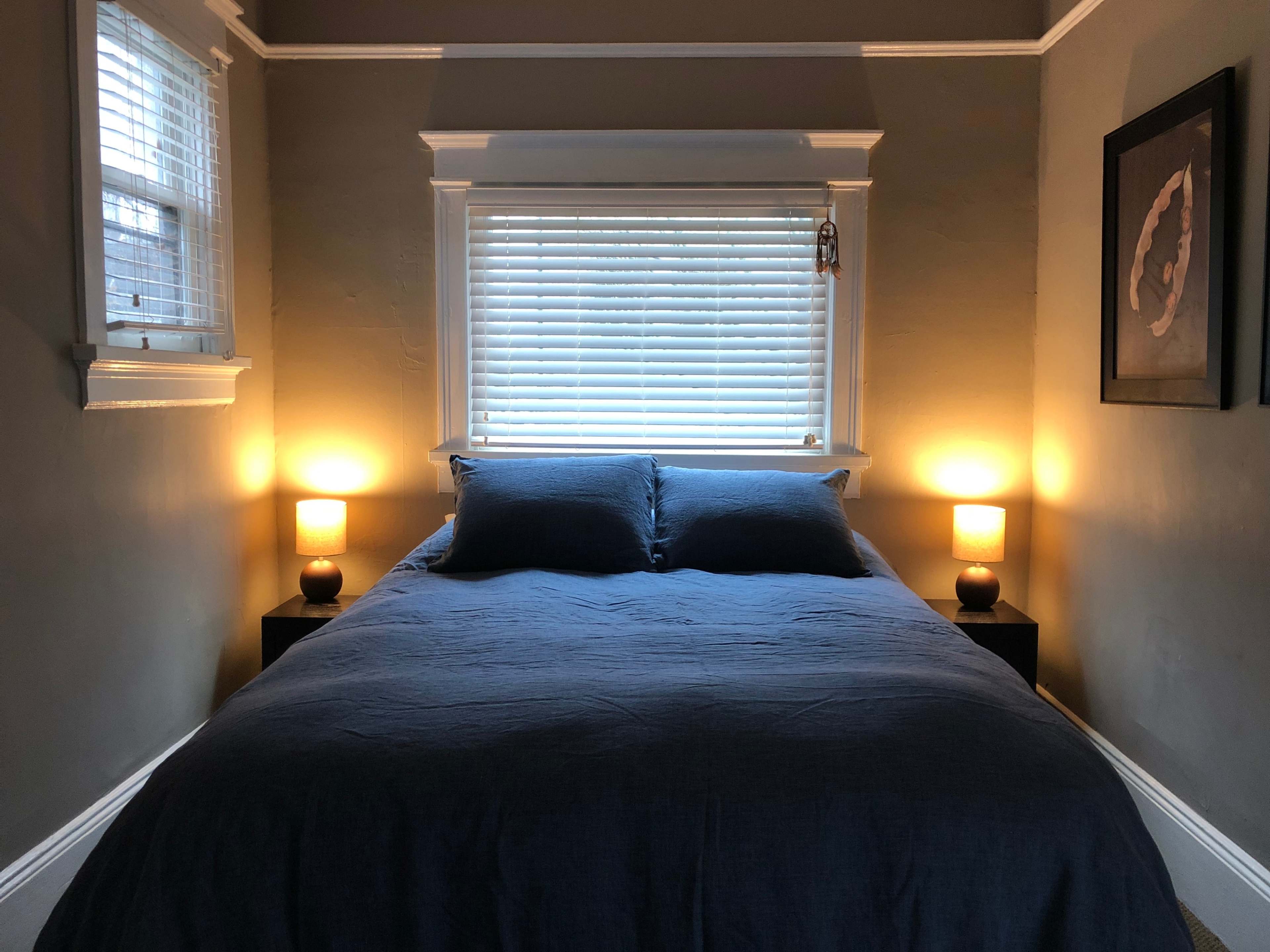 A neatly made bed with blue bedding is positioned between two bedside tables with illuminated lamps beneath a window with closed blinds.