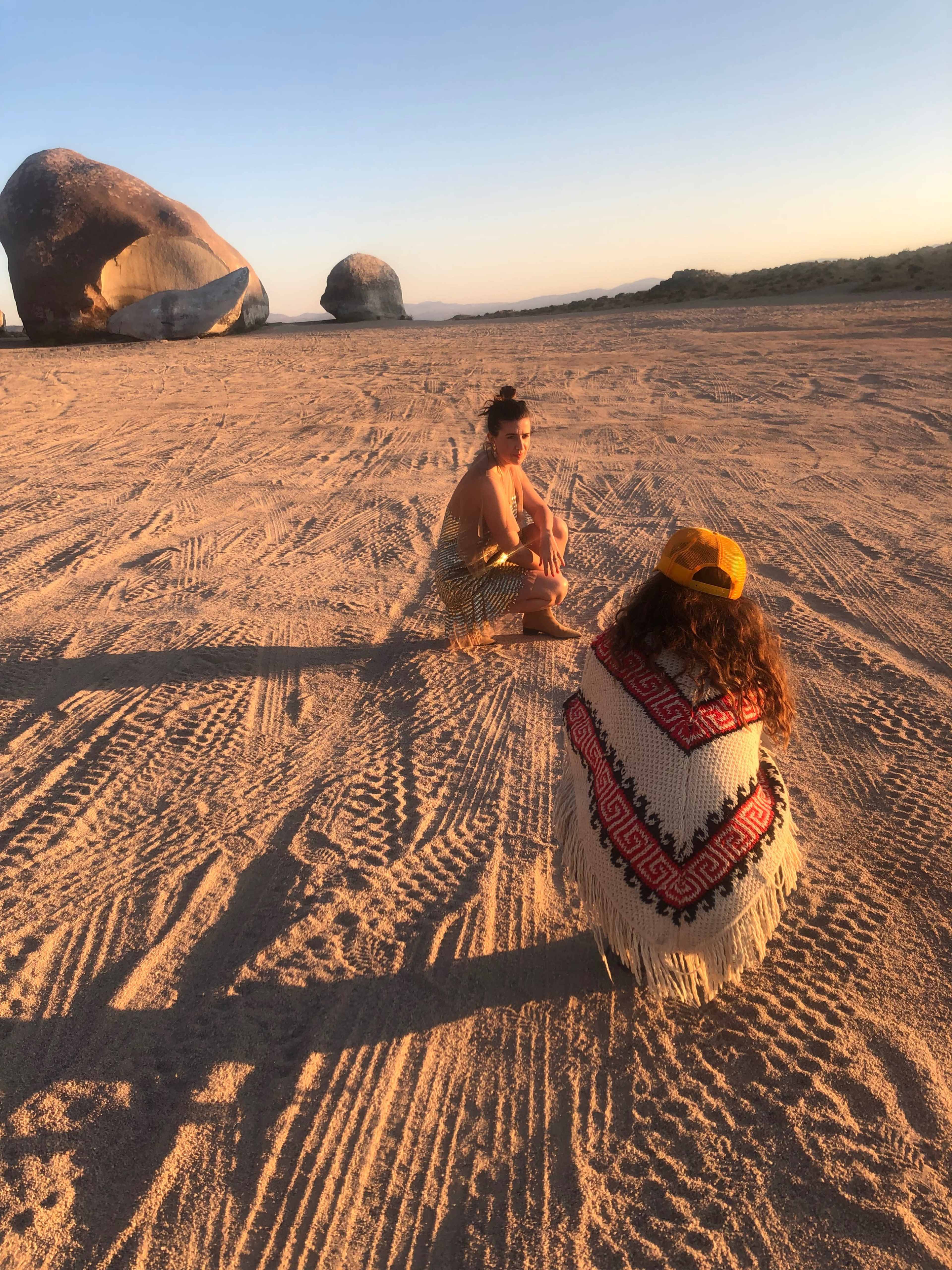 Two individuals are sitting on a sandy terrain with large rock formations in the background during sunset.