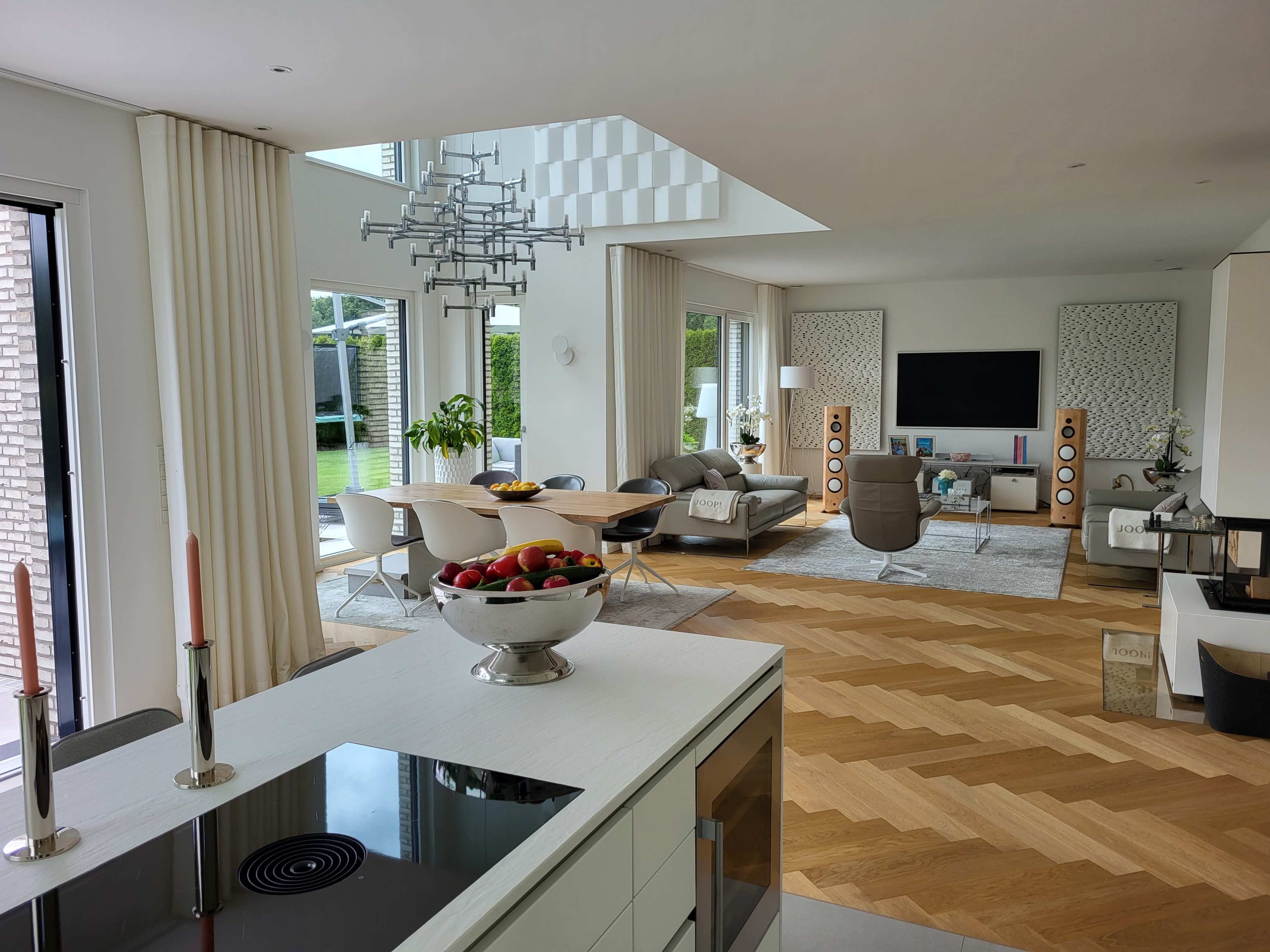 A modern living space featuring a kitchen area with a sleek stove, a dining table with a bowl of fruit, and a lounge area furnished with a gray sofa and large television.