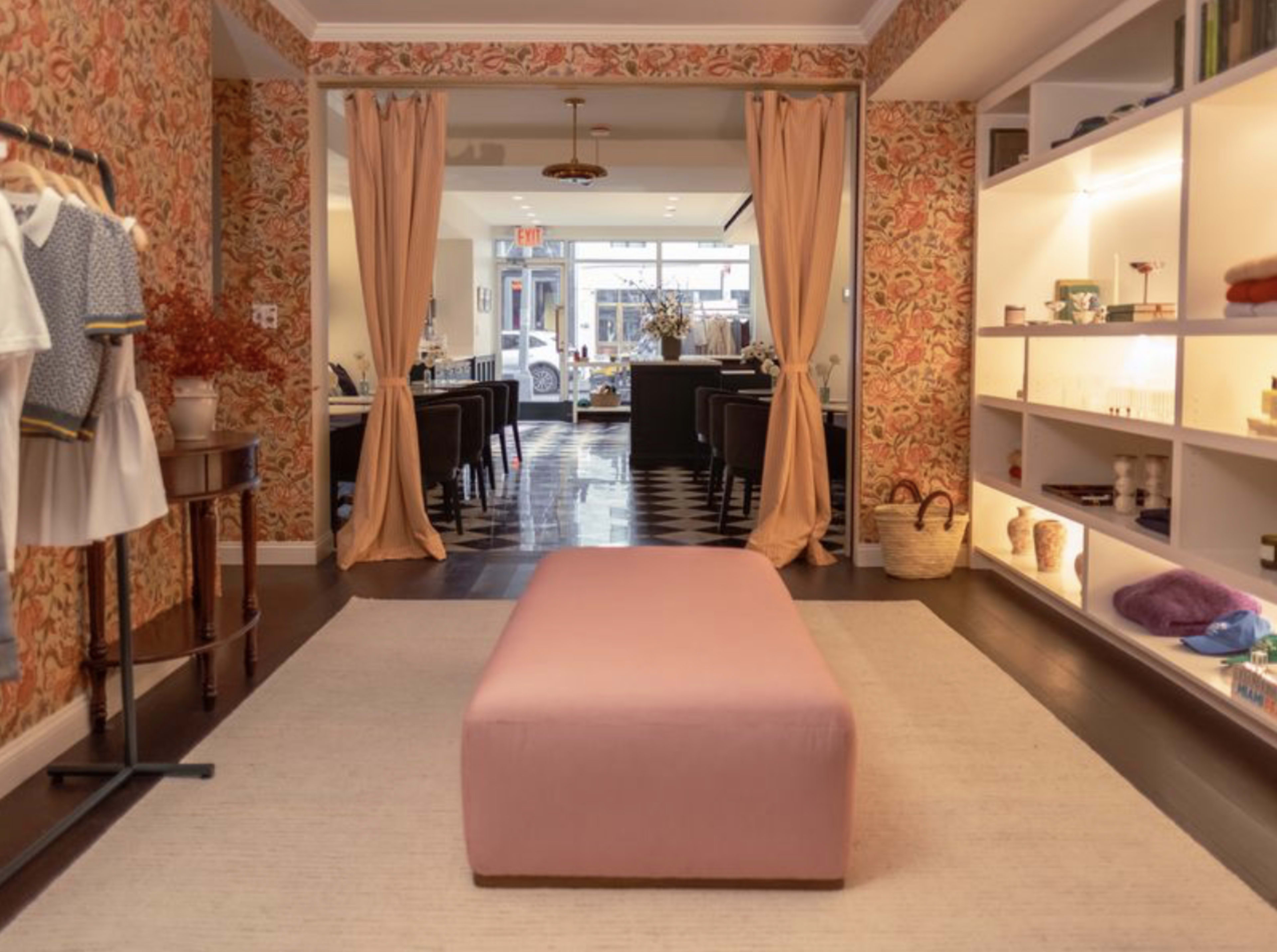 The image shows the interior of a boutique with floral wallpaper, a pink upholstered bench in the center, and a view into a seating area with tables and chairs.