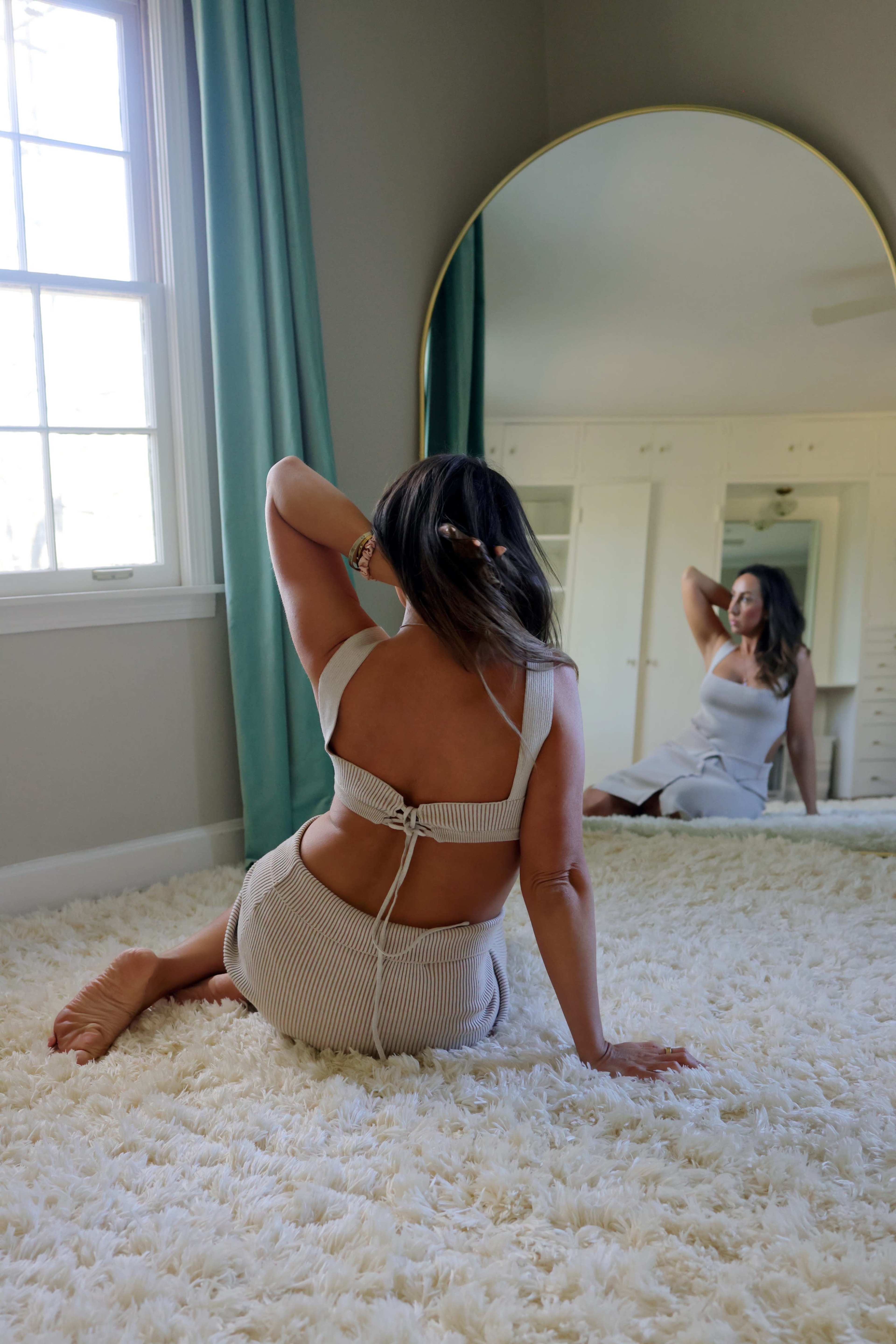 A woman poses on a plush rug in front of a mirror, showcasing a backless outfit while sitting on the floor.