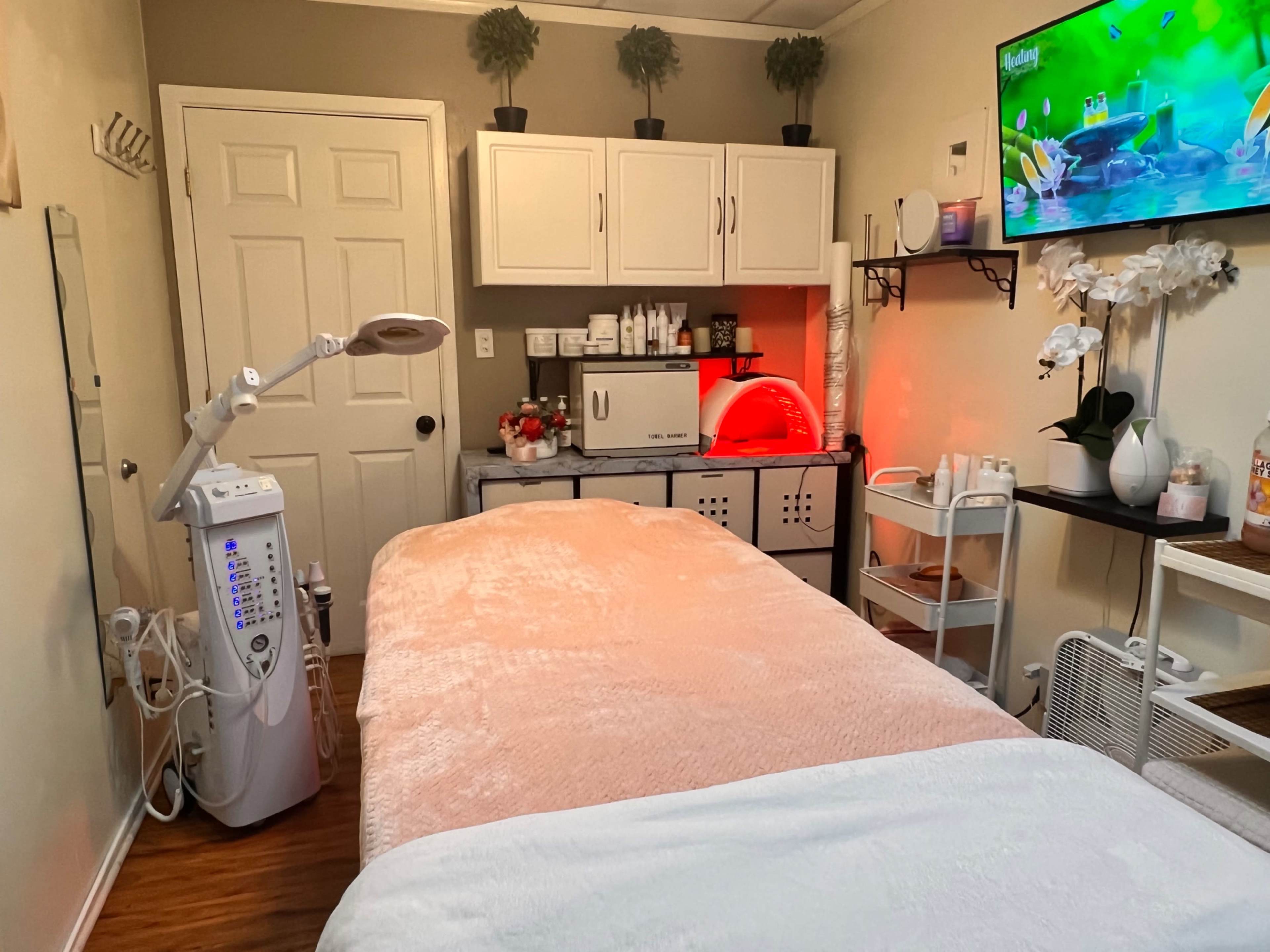 The image shows a tranquil treatment room featuring a massage table, a variety of skincare products on shelves, and a wall-mounted television displaying a serene scene.