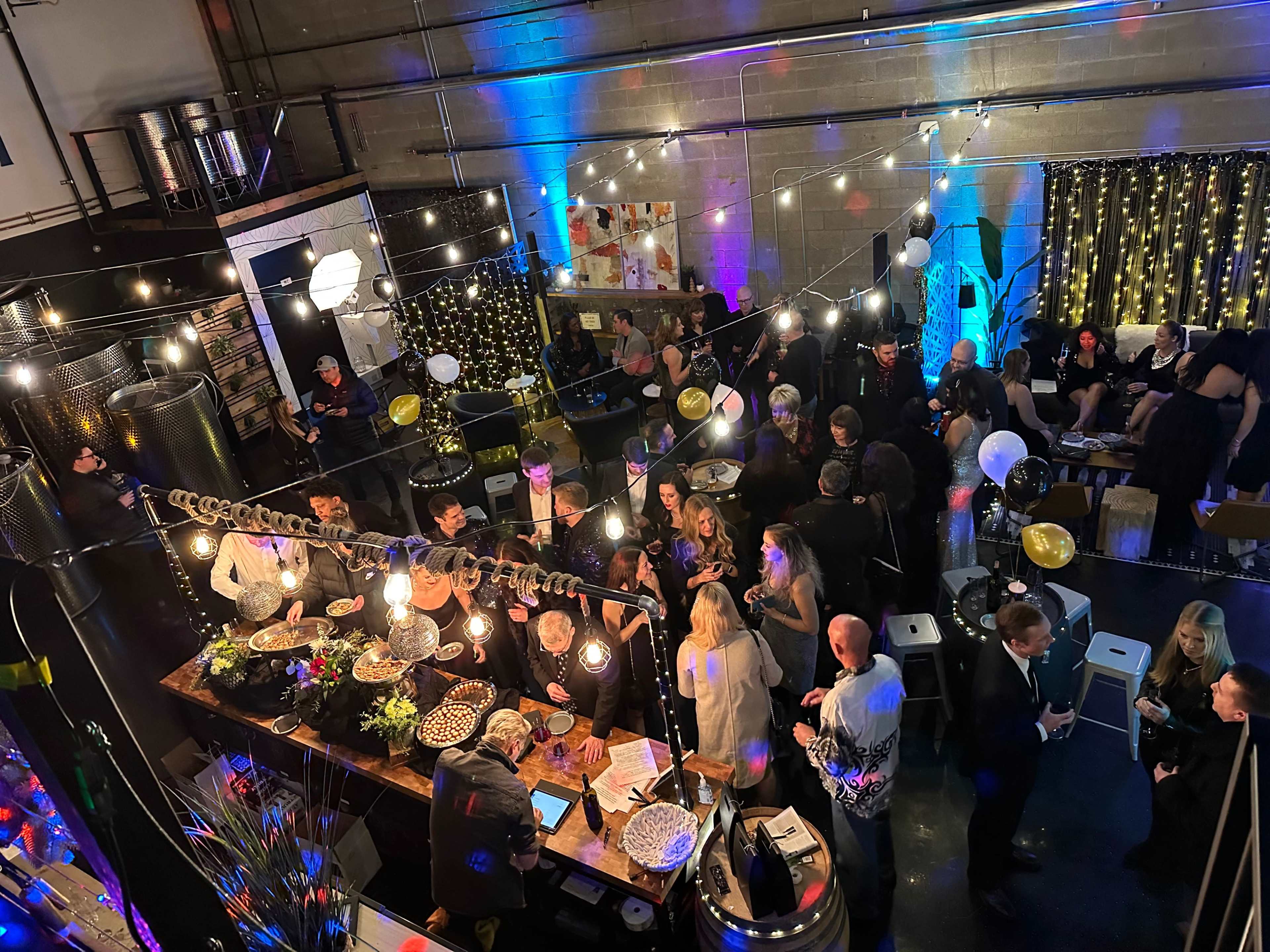 A lively indoor event with people socializing around a bar adorned with decorative lights and tables, set amidst a backdrop of balloons and festive decorations.
