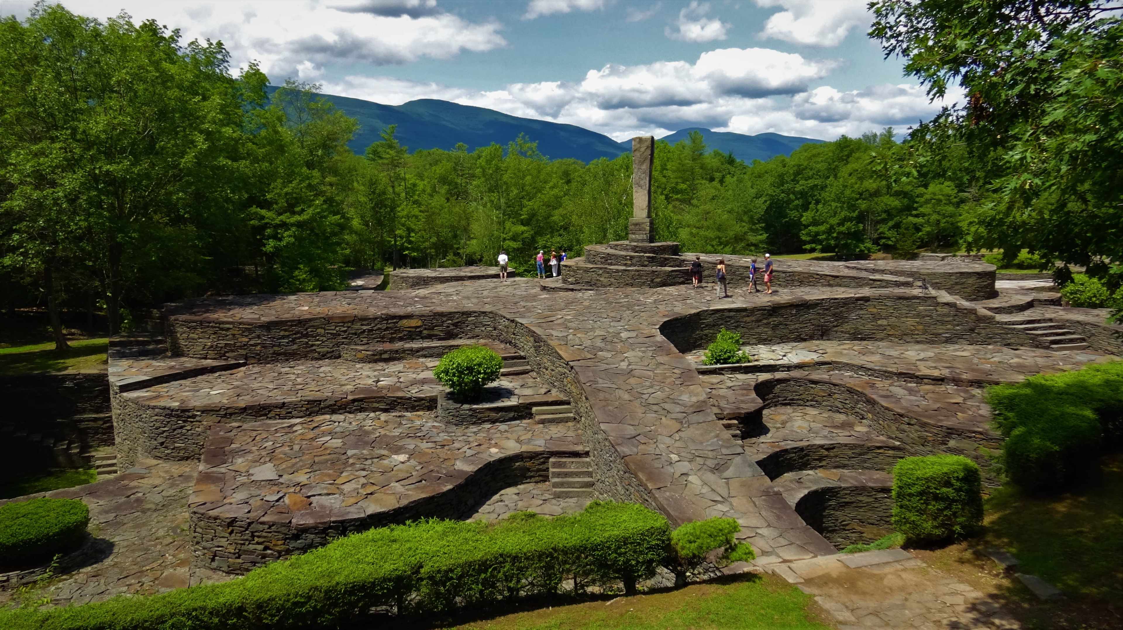 An exposed stone structure surrounded by trees and visitors, with a tall monument in the center.