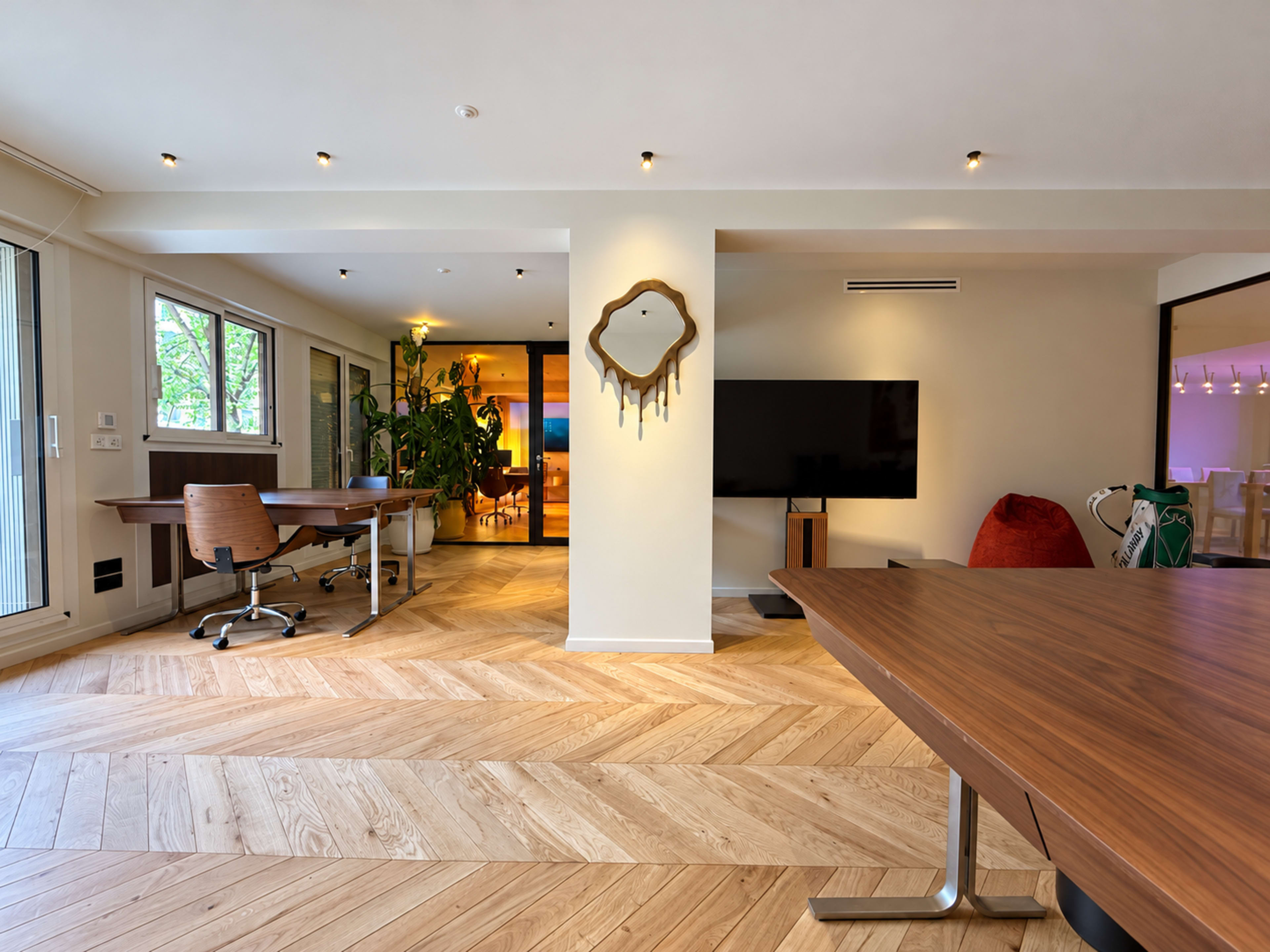 The image shows a modern open-plan office space with wooden flooring, a large flat-screen TV on the wall, and a mix of workstations and casual seating.