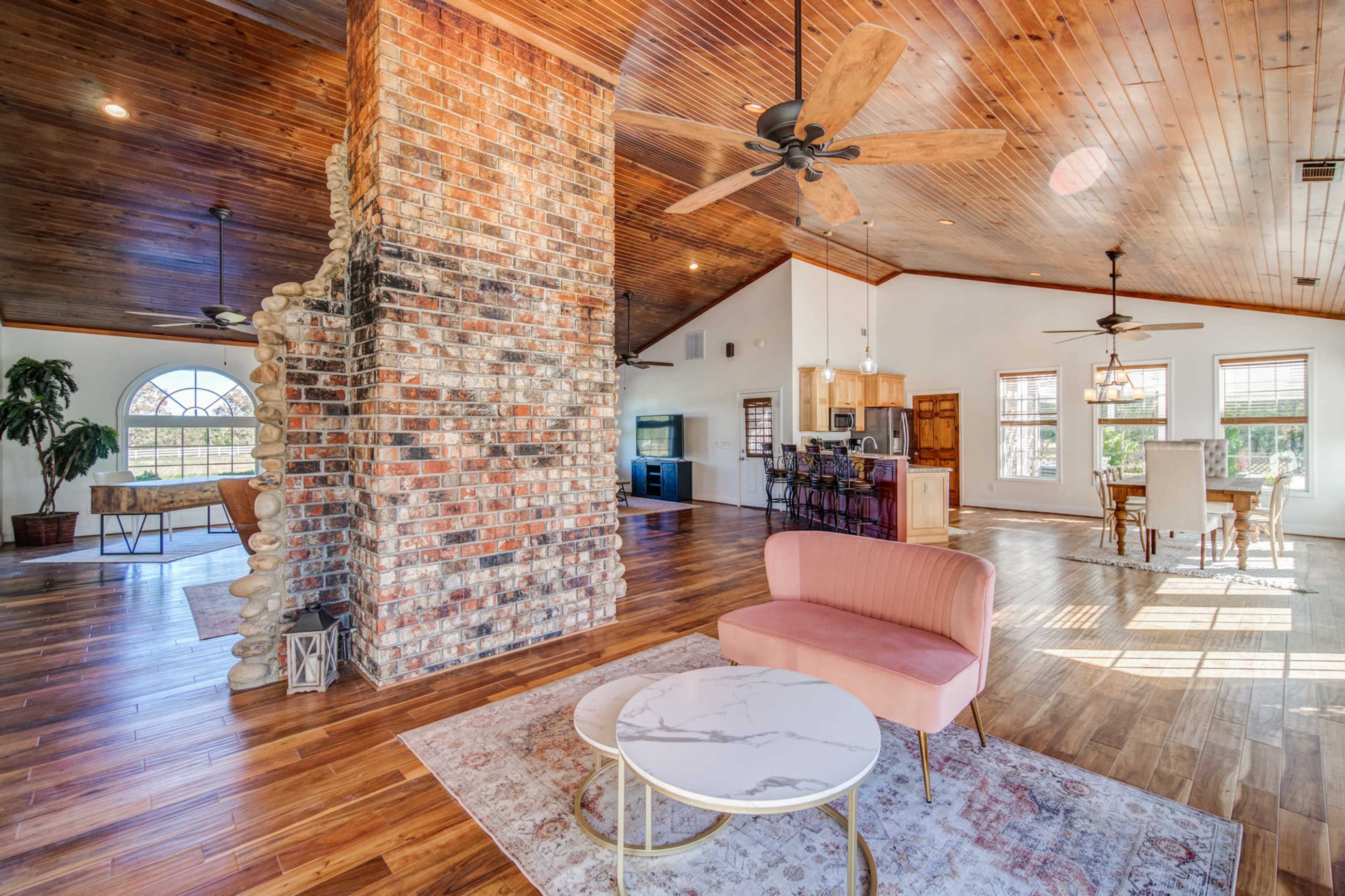 The image shows a spacious living area featuring a brick fireplace, a pink sofa, round tables, and wooden flooring, with large windows allowing natural light.