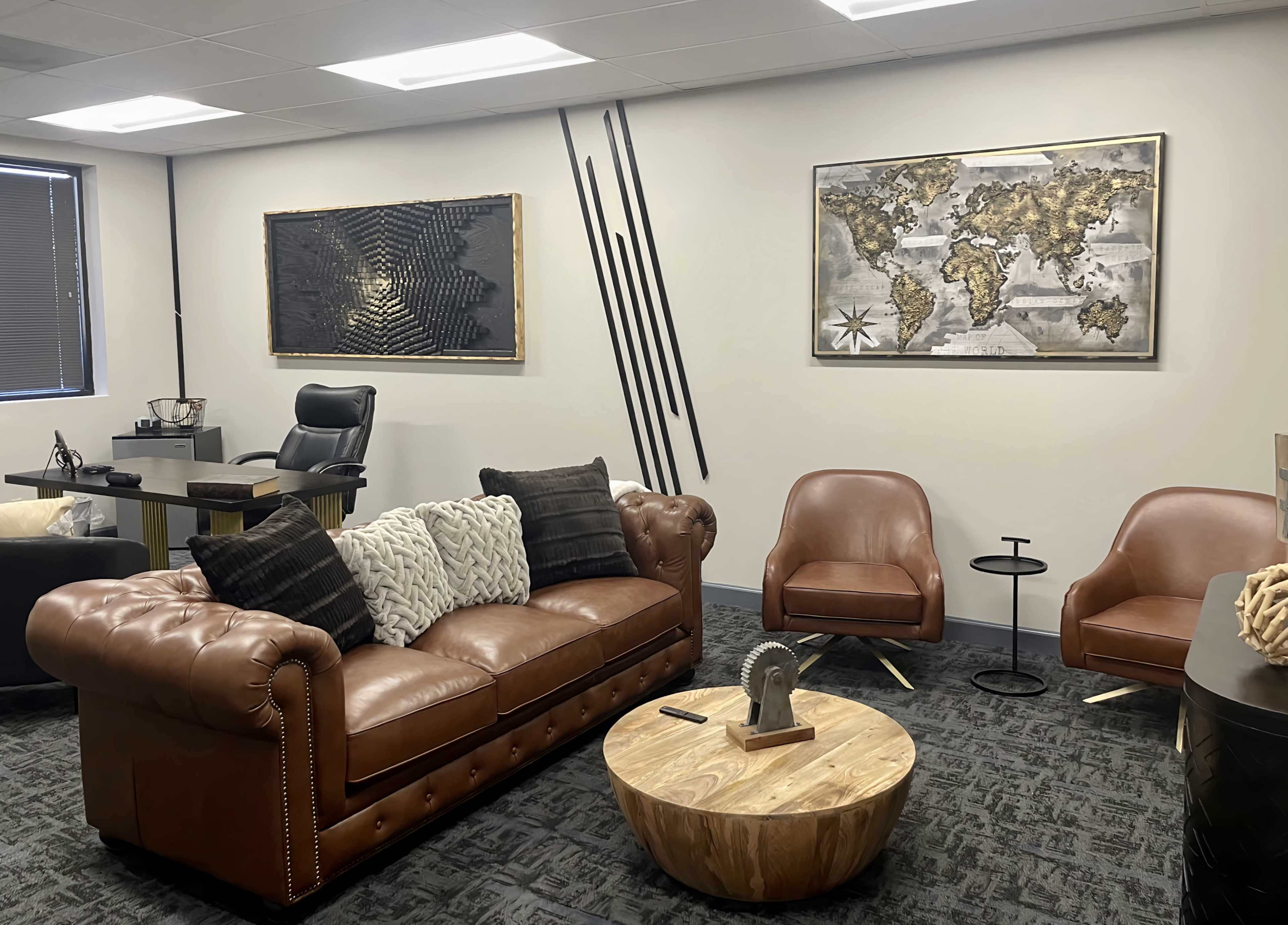 A modern office space featuring a brown leather sofa with decorative pillows, a round wooden coffee table, two stylish chairs, and wall art including a world map and abstract designs.