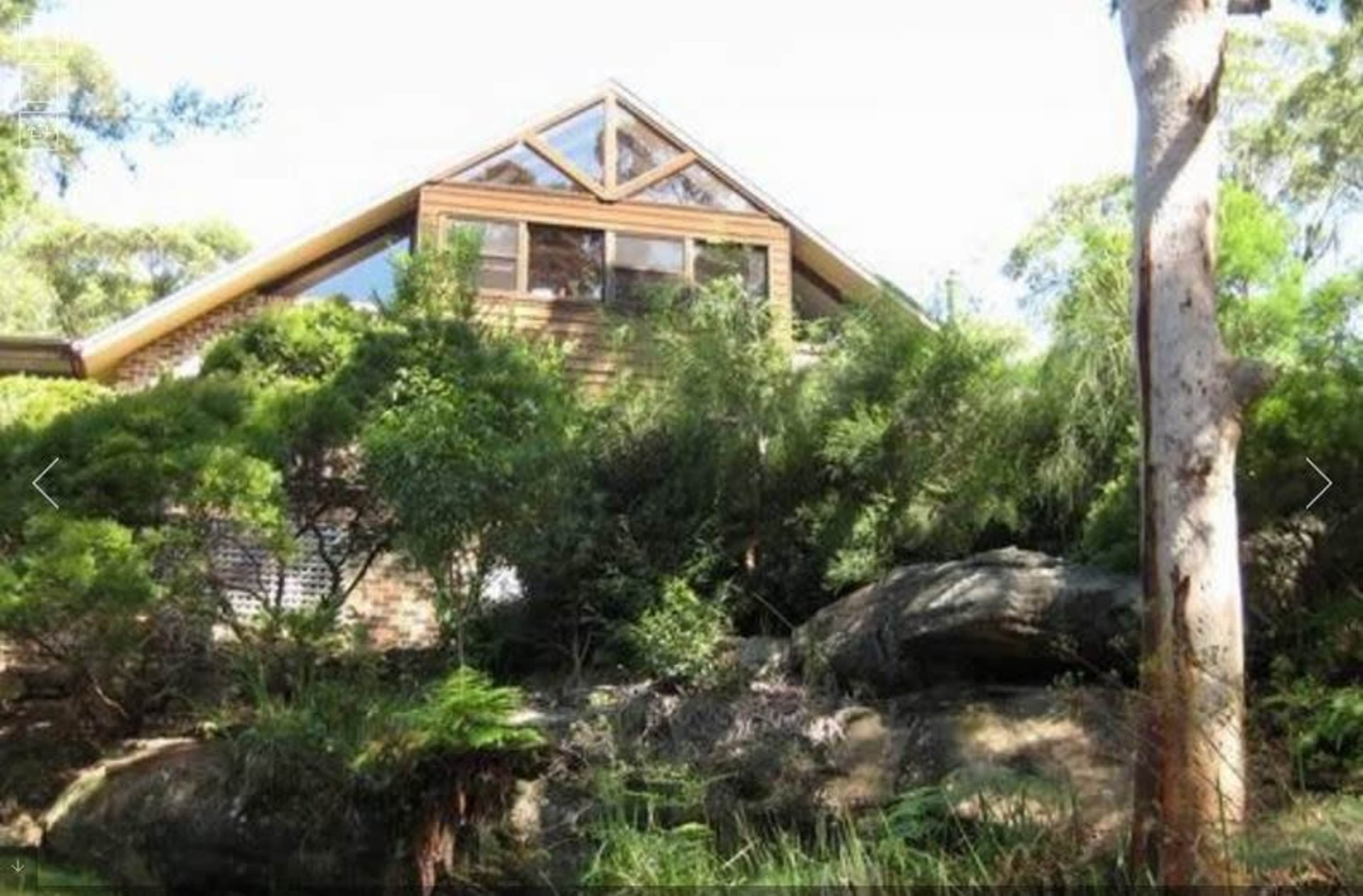A modern house with a triangular roof and large windows sits amid lush greenery and rocks.