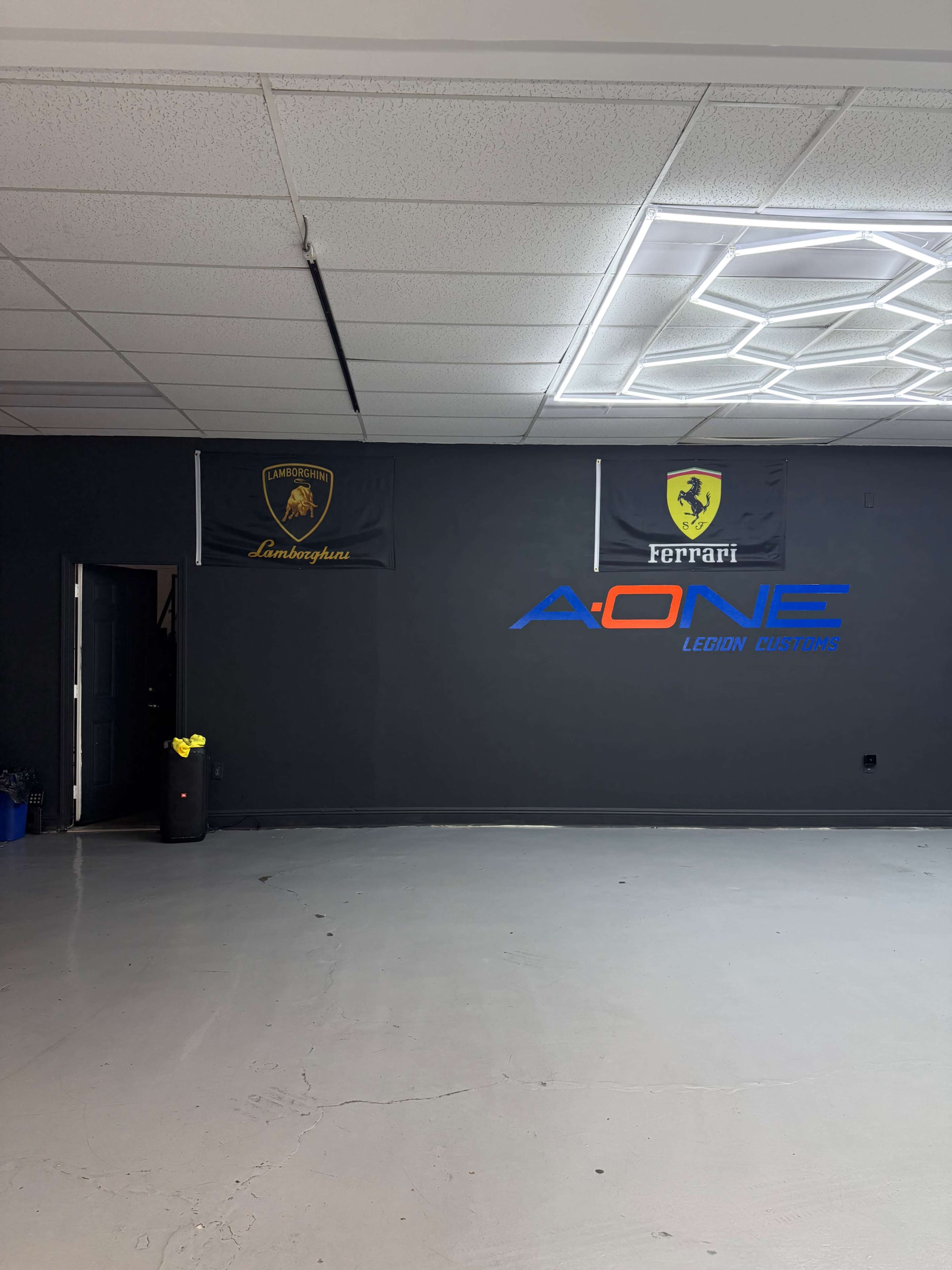 The image shows an interior space with a gray floor, black walls featuring flags of Lamborghini and Ferrari, and a prominent blue logo of A-ONE Legion Customs on the wall.