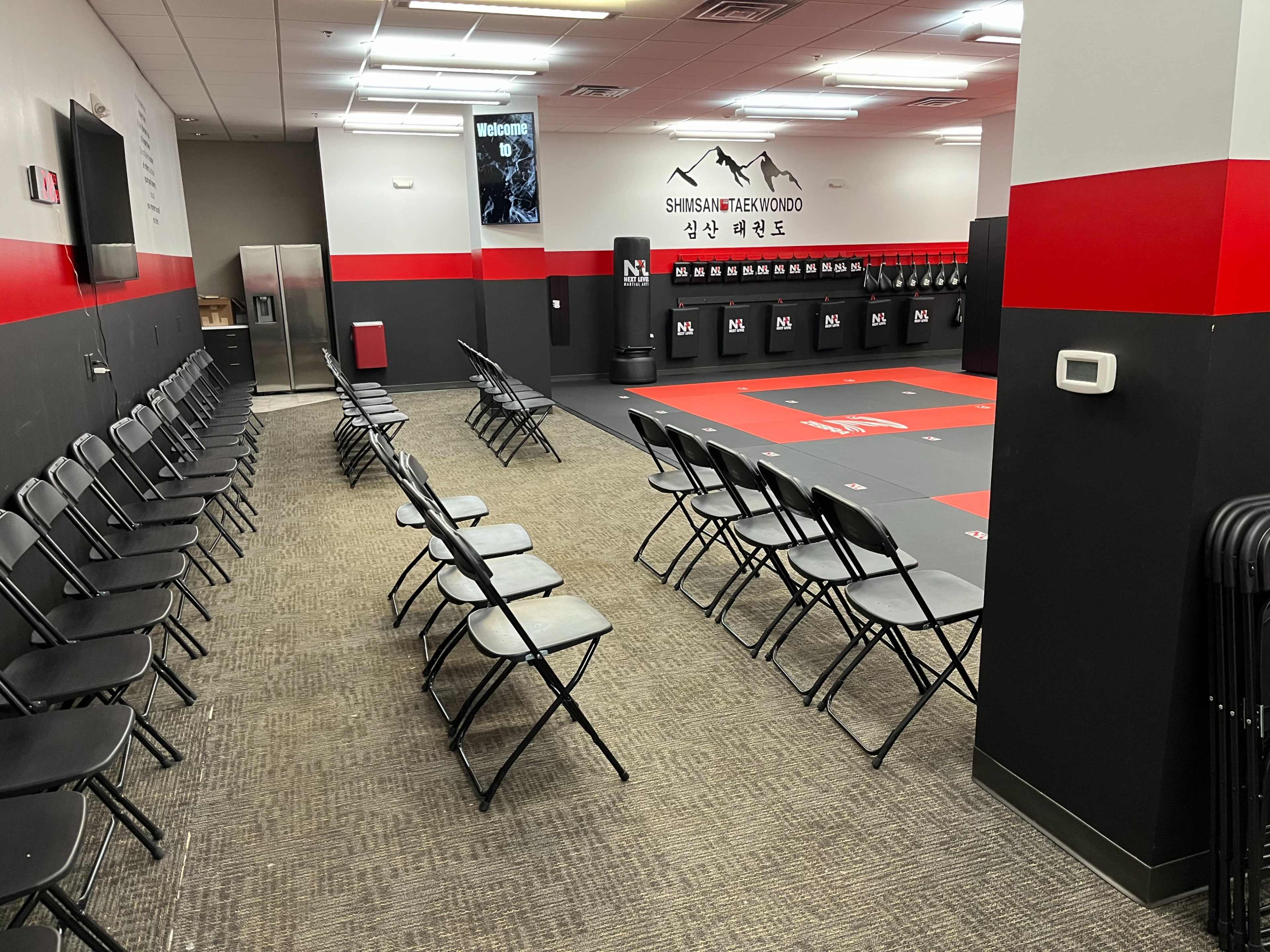 The image shows a training room with rows of folding chairs arranged beside a red and black wrestling mat, and a wall of lockers in the background.