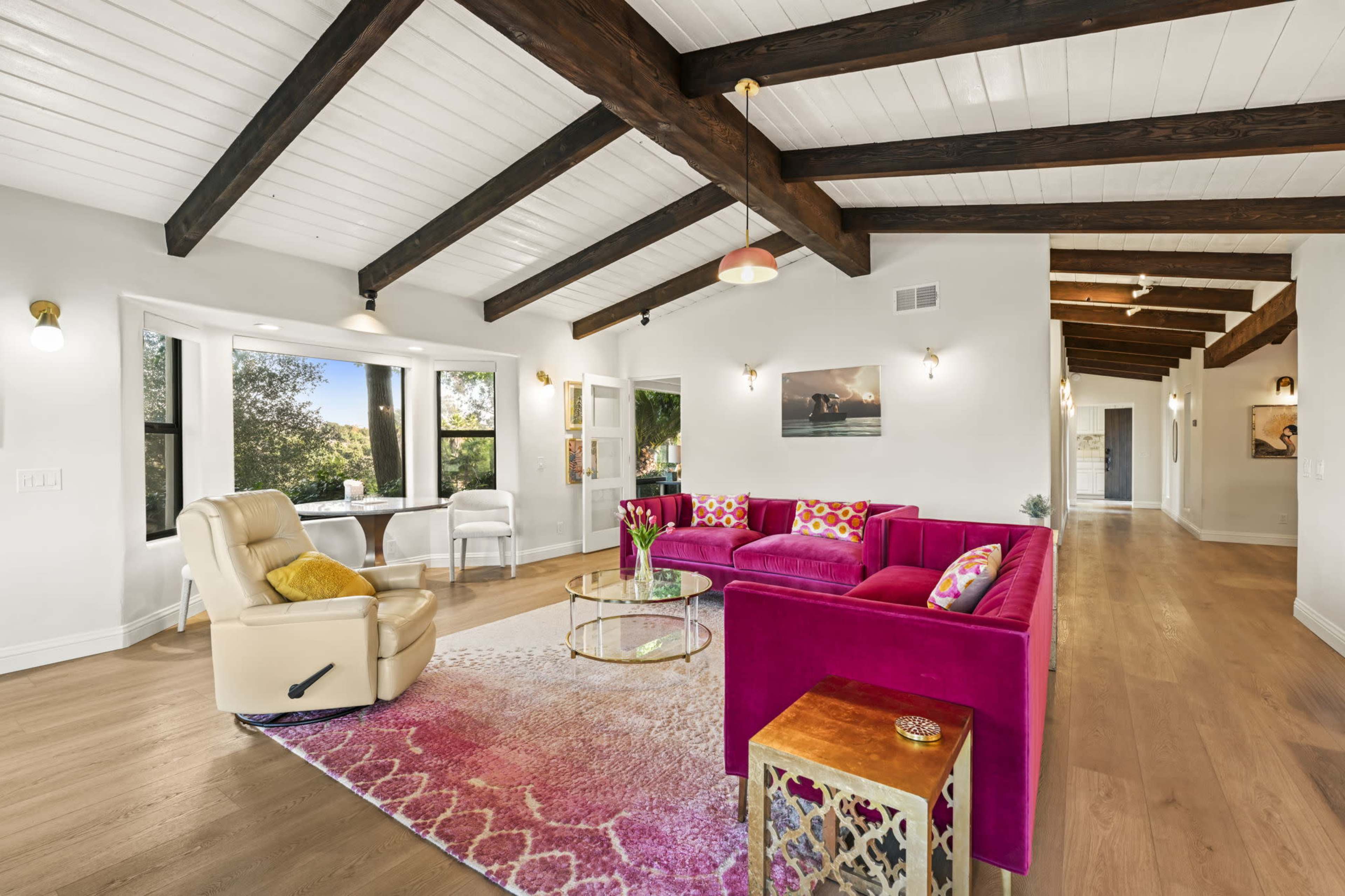 The image shows a bright living room featuring a pink sofa, a round coffee table, and a reclining chair, with exposed wooden beams on the ceiling and large windows letting in natural light.