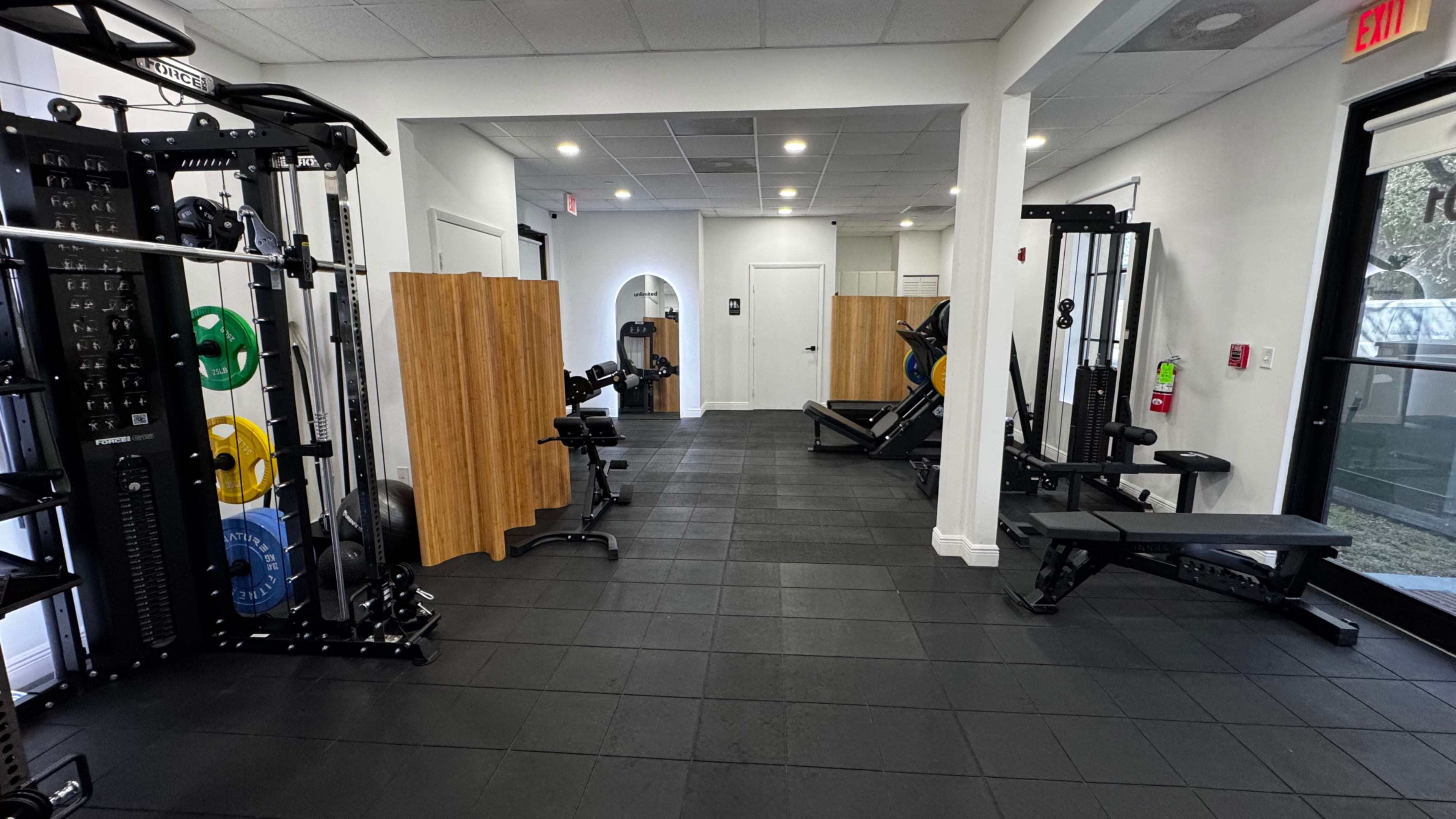The image shows a fitness facility with various workout equipment, including weights, benches, and exercise machines, arranged in an open layout.