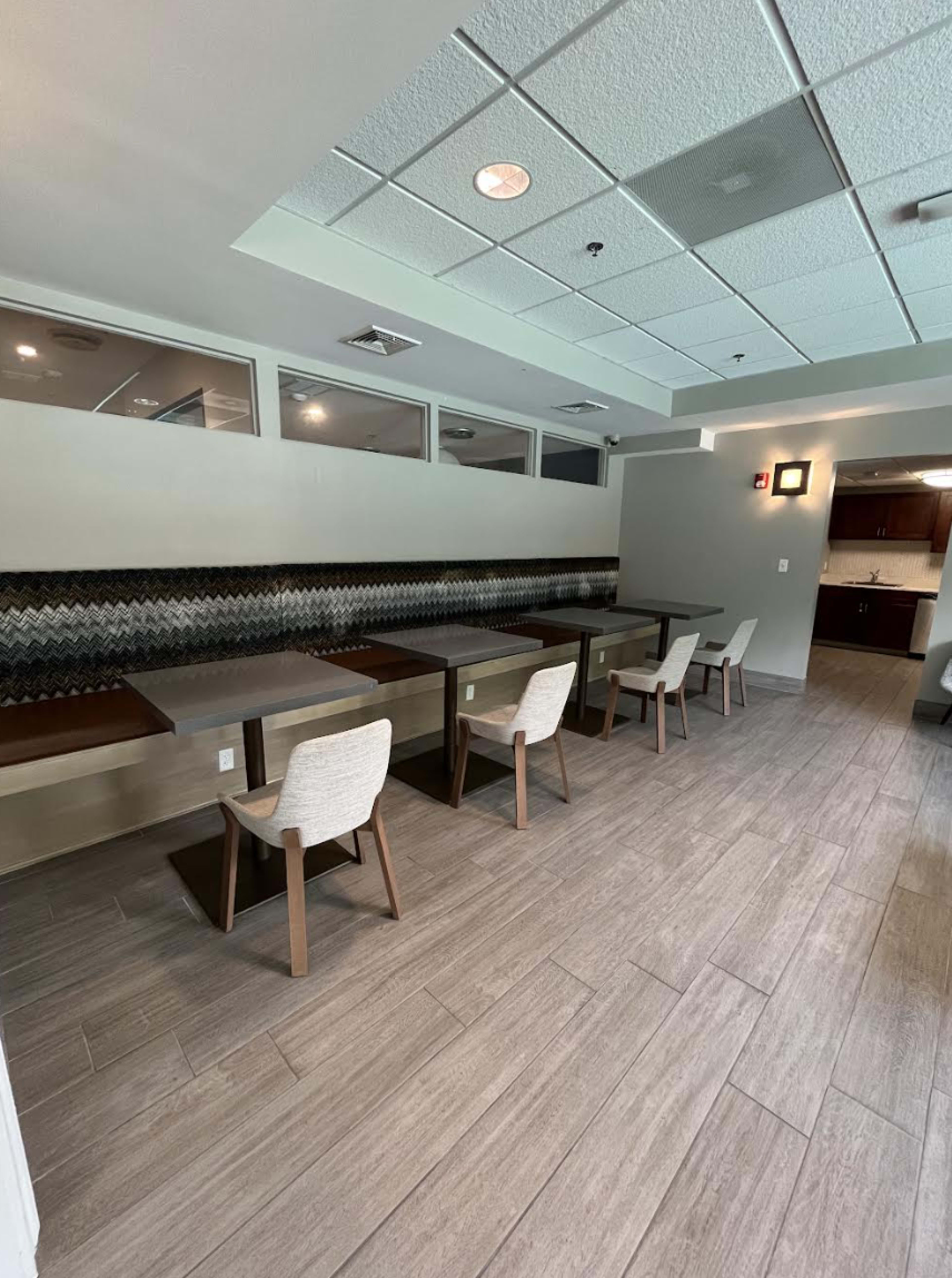 The image shows a modern dining area with several tables and chairs arranged in a spacious, well-lit room with neutral-colored walls and wood-style flooring.