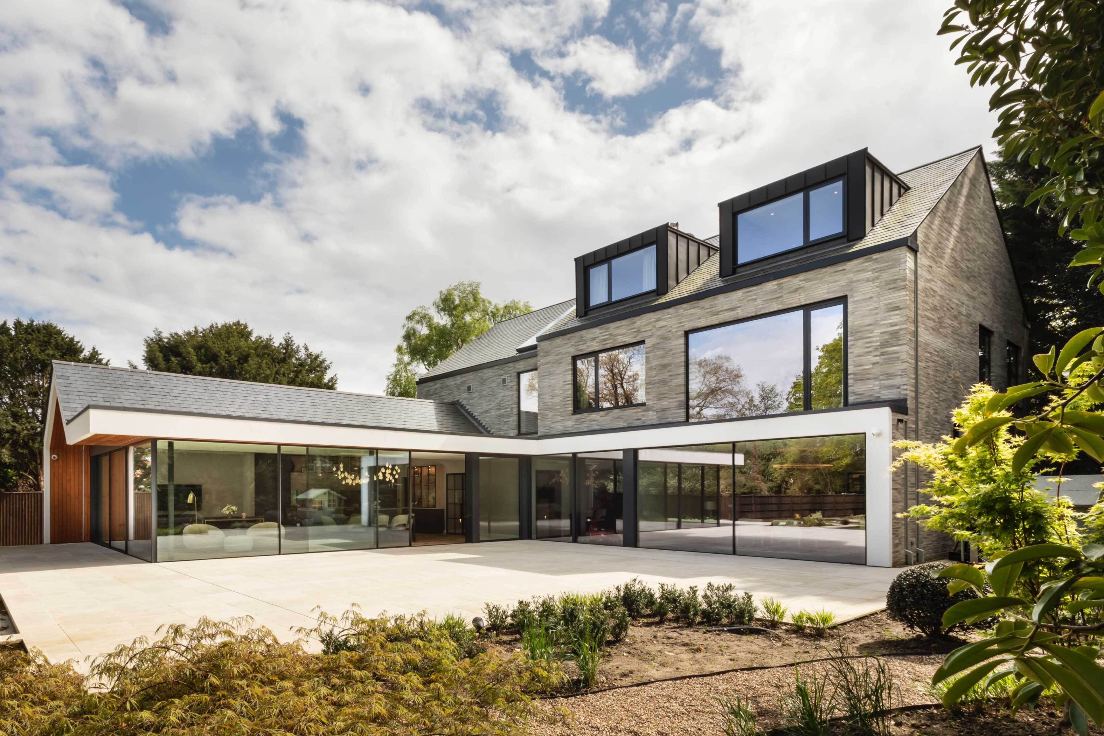 A modern two-story home with large glass windows, a flat roof section, and a stone exterior surrounded by landscaped greenery.
