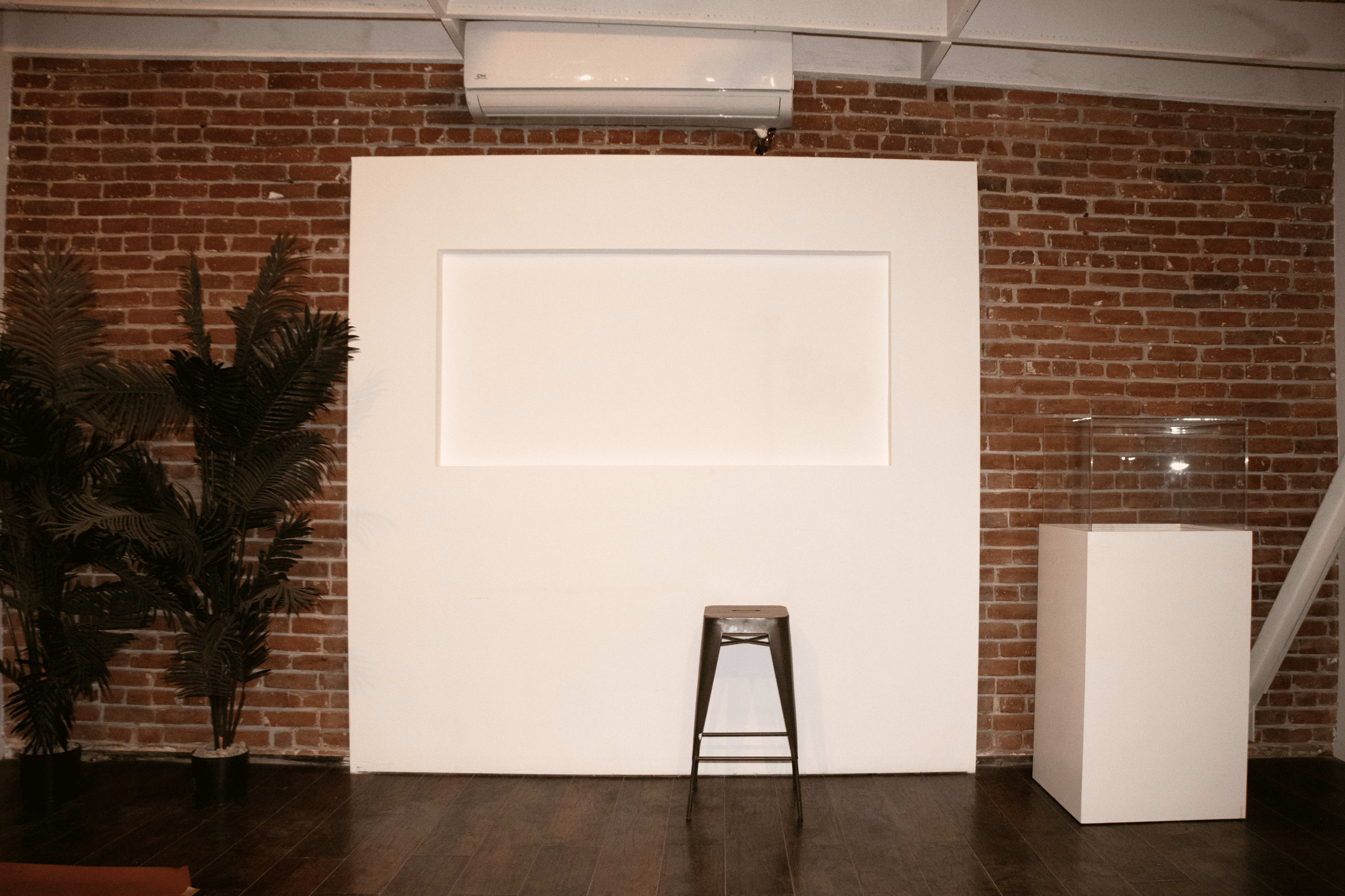 A minimalist interior with a brick wall, a blank white panel, a metal stool, and a glass display case beside a potted plant.