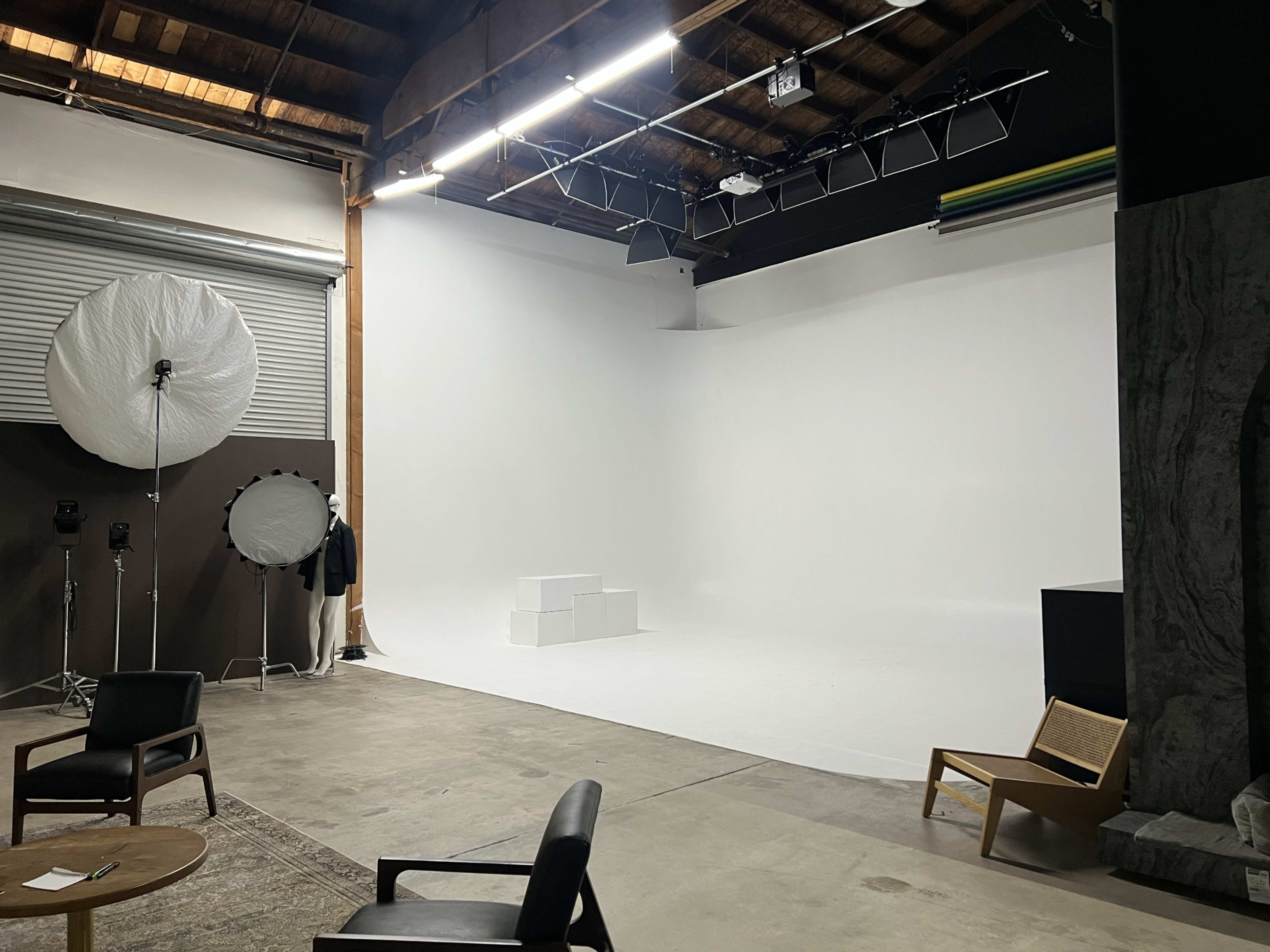A spacious photography studio features a blank white backdrop, scattered lighting equipment, and minimalistic furniture.