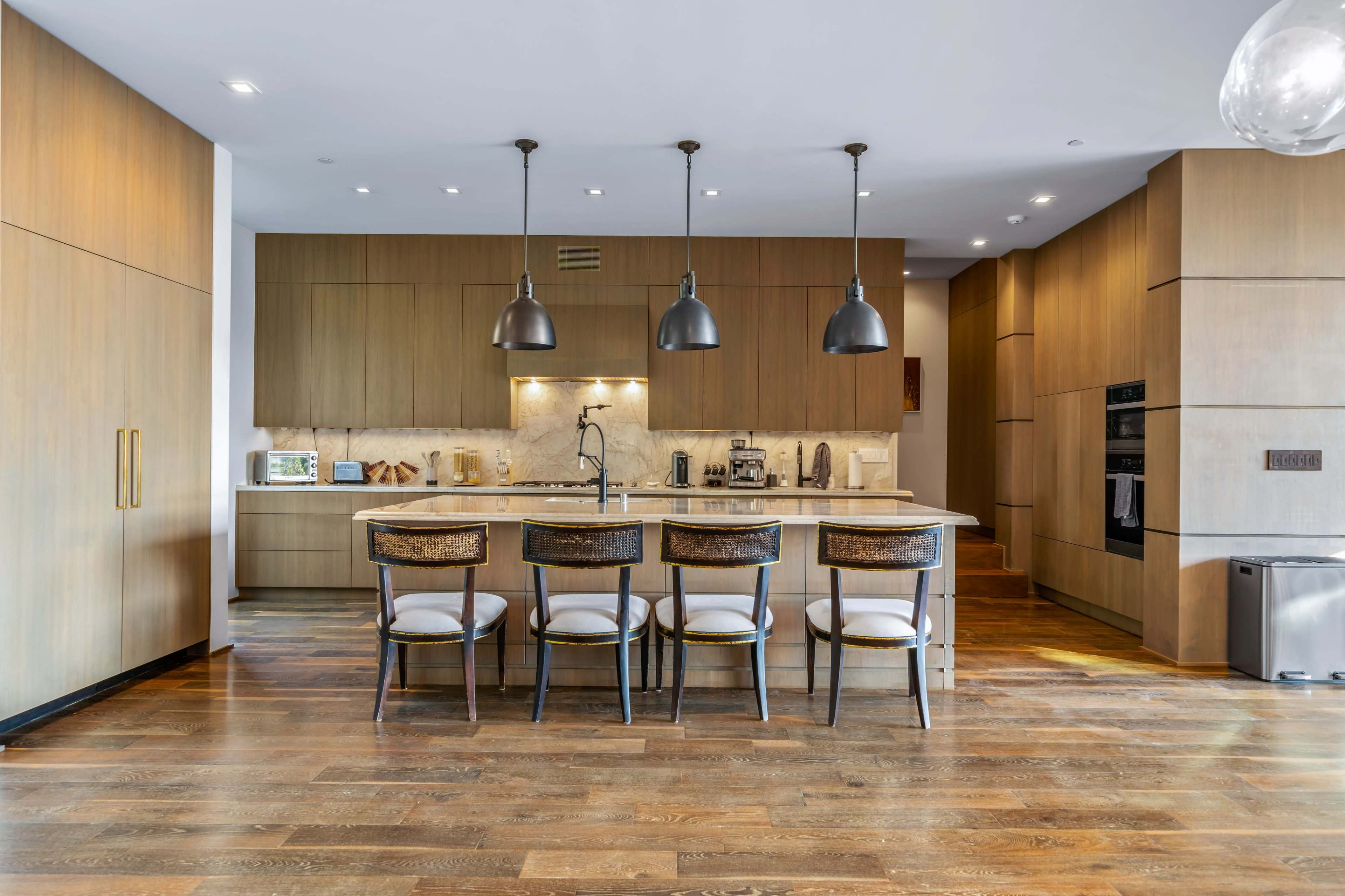 A modern kitchen features wooden cabinetry, a large island with seating, and pendant lights above.