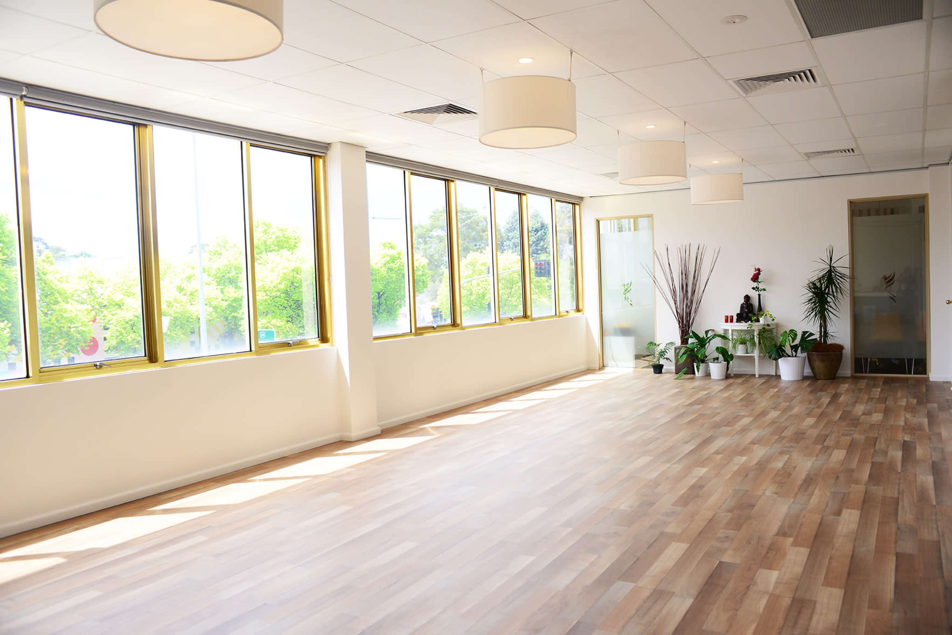 A bright, spacious room with large windows, wooden floors, and potted plants along the walls.