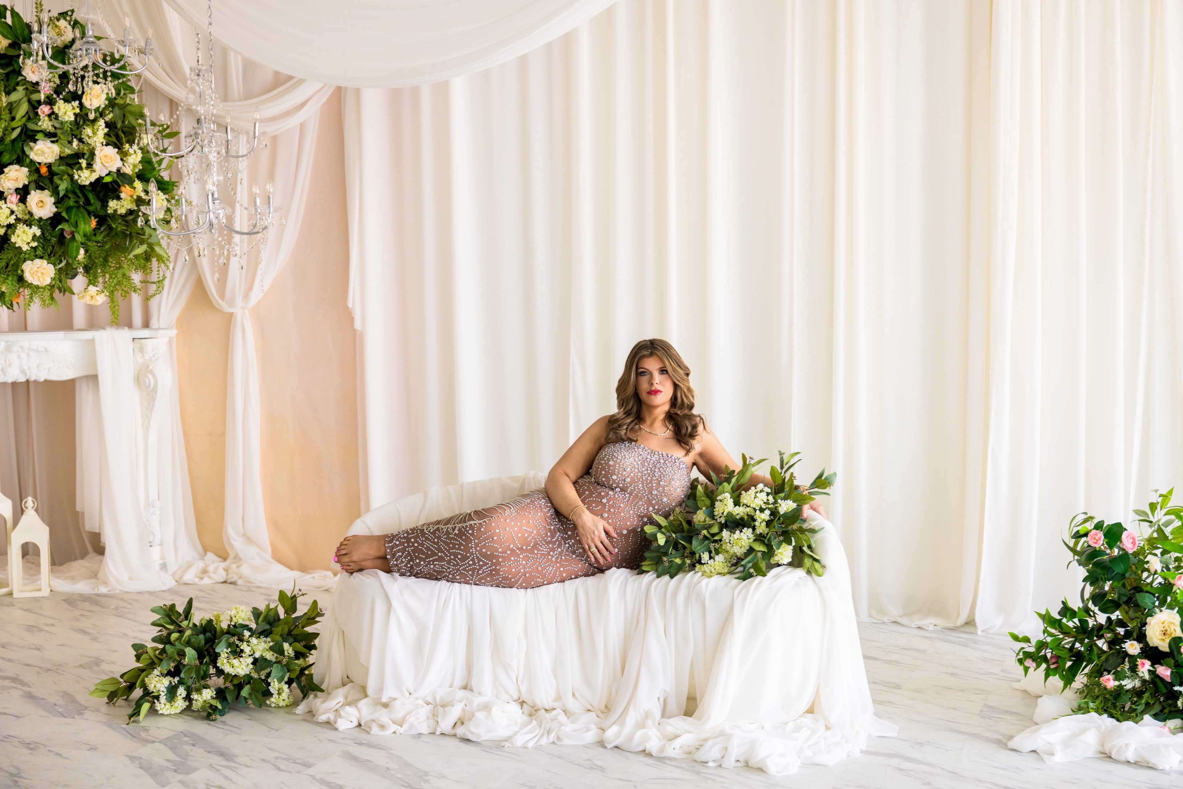 A woman in a sequined gown reclines on a white sofa surrounded by floral arrangements in a decorated indoor setting.