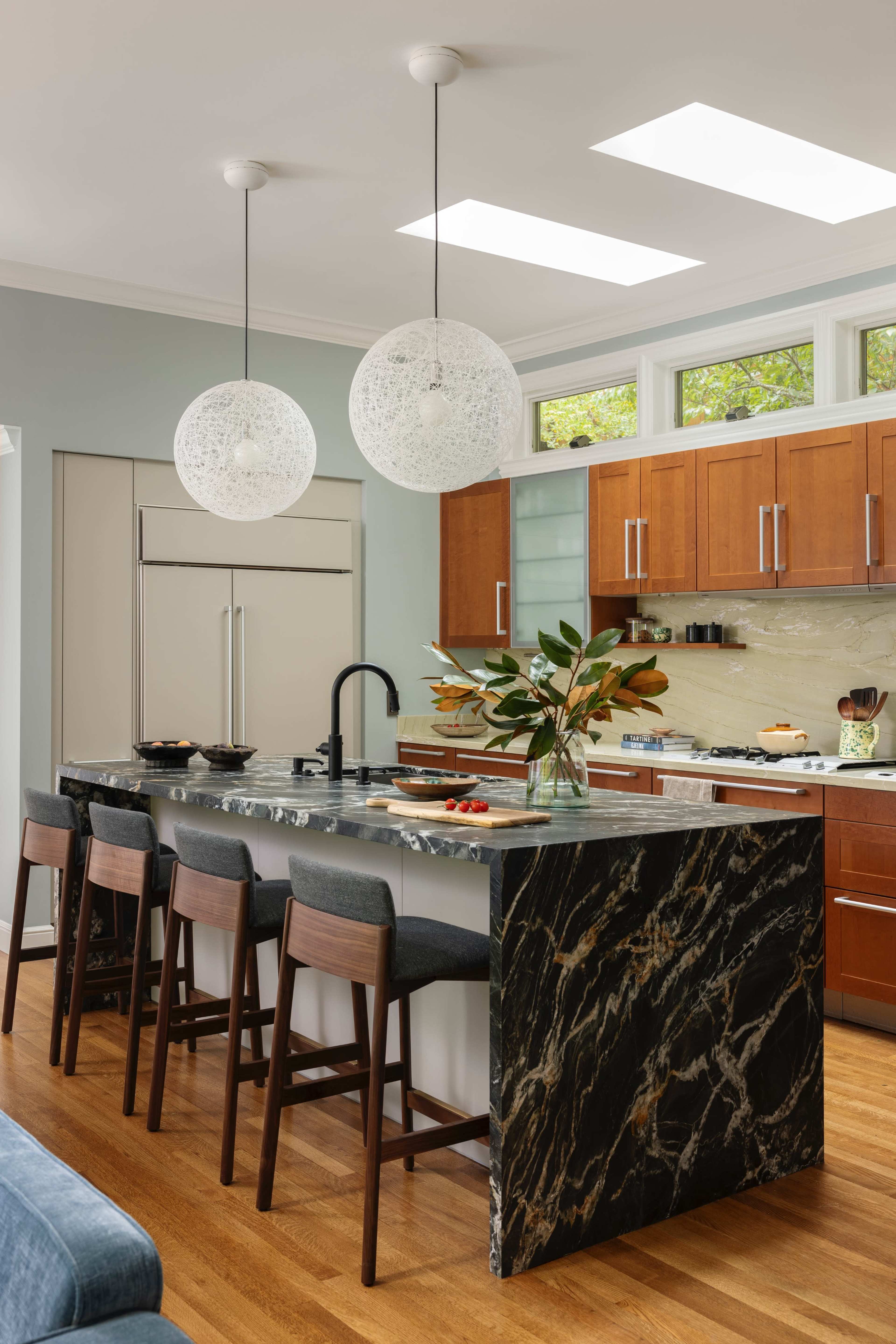 A modern kitchen features a marble island with seating, wooden cabinetry, and pendant lights above the countertop.