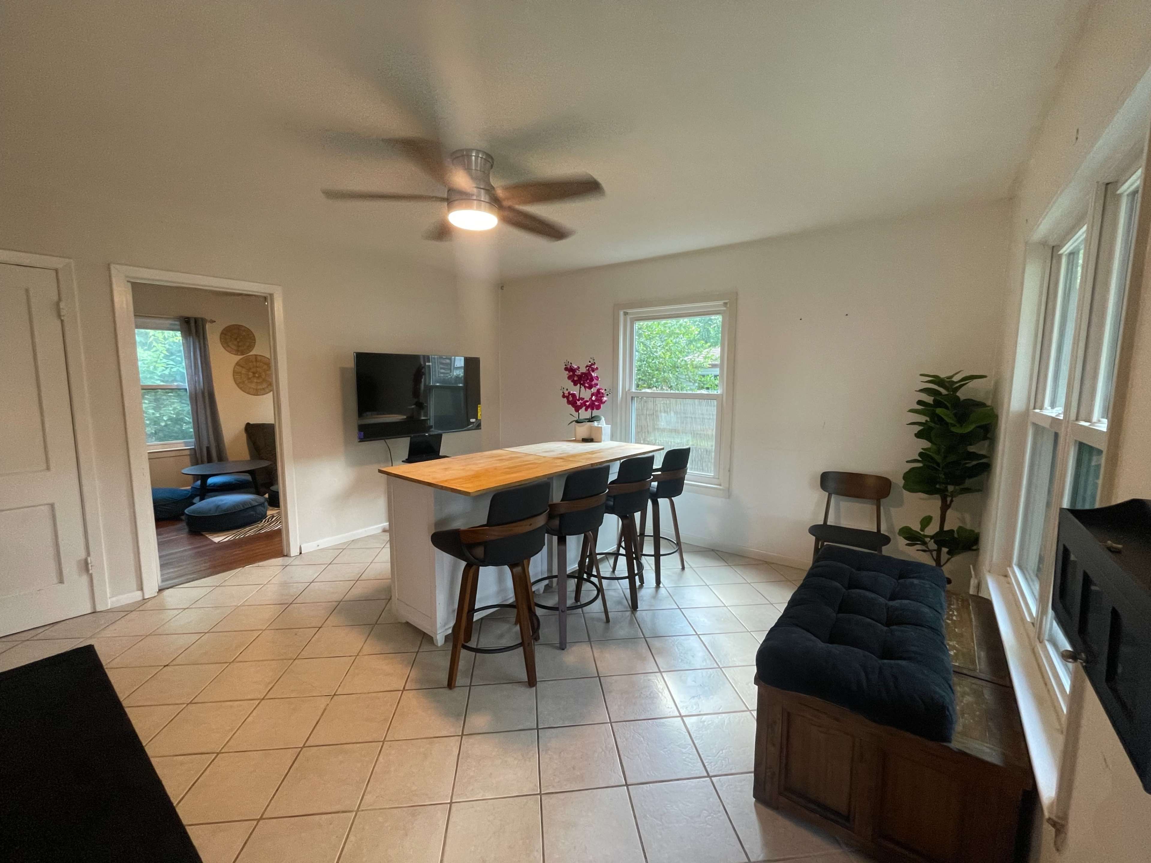 The image shows a bright, cozy living space with a kitchen island, four wooden barstools, a television mounted on the wall, and large windows allowing natural light to enter.