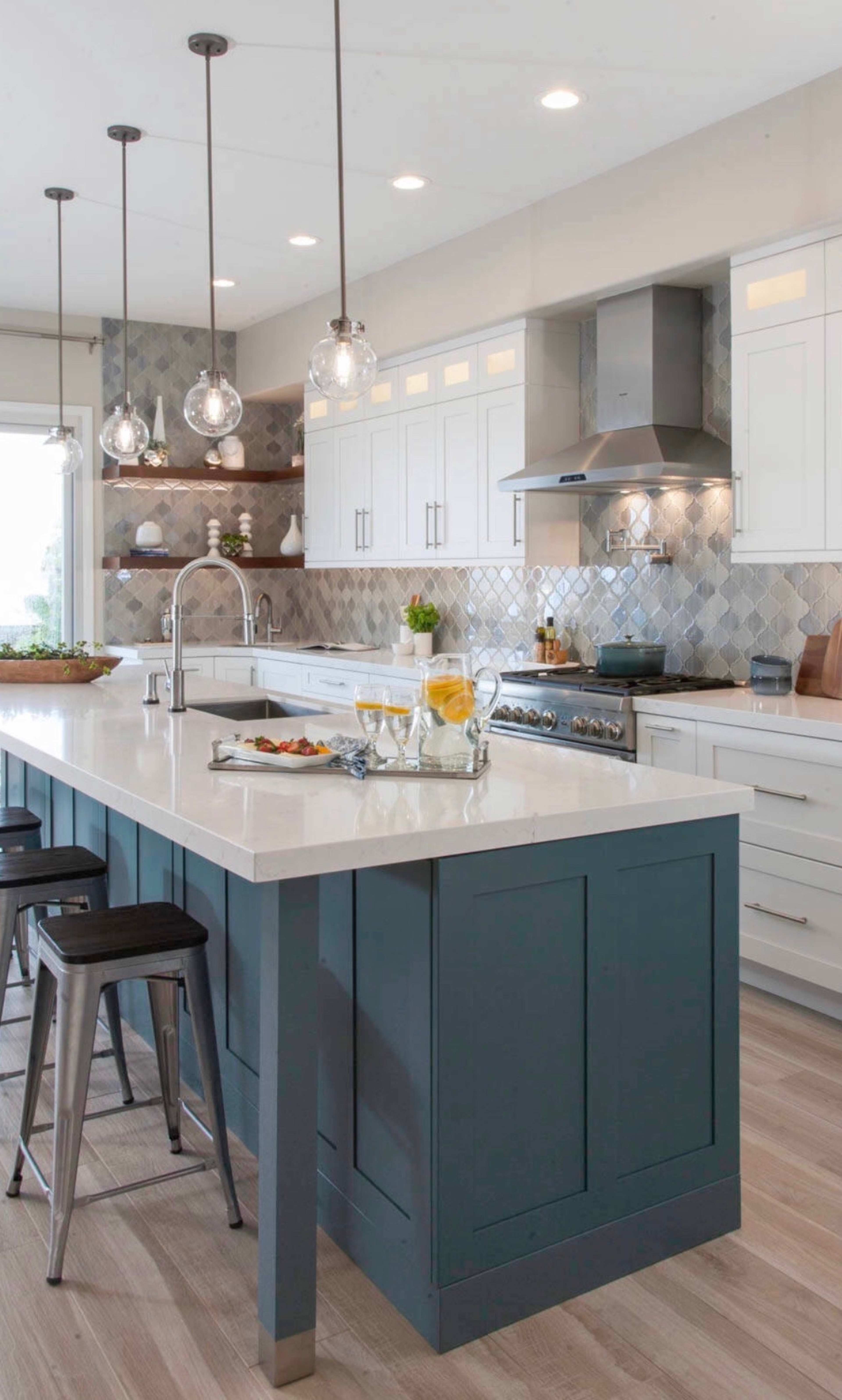 A modern kitchen features a large island with a dark blue base, white cabinets, and stainless steel appliances, illuminated by pendant lights.
