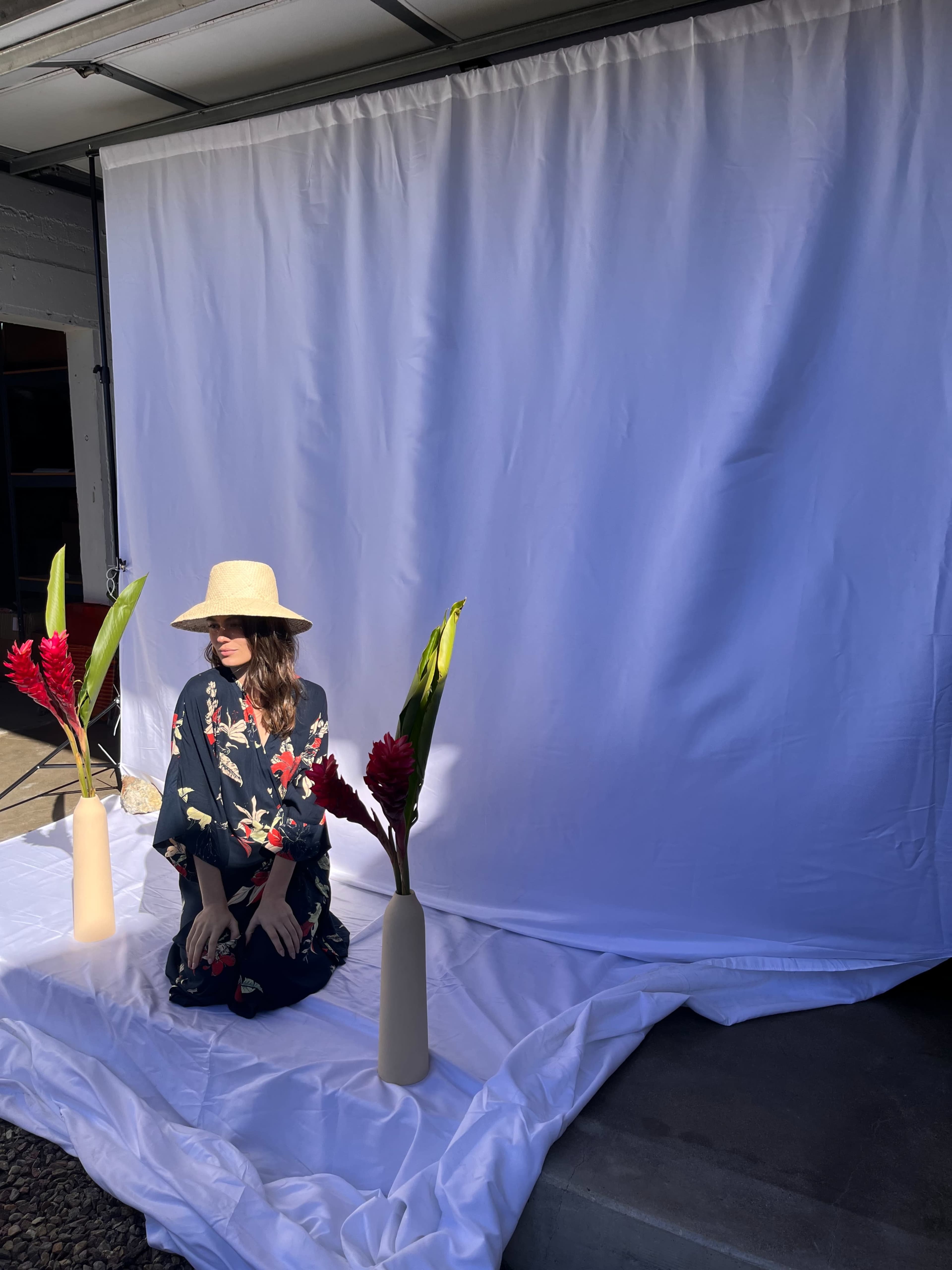 A woman in a floral outfit sits in front of a white backdrop with two vases containing flowers on either side.