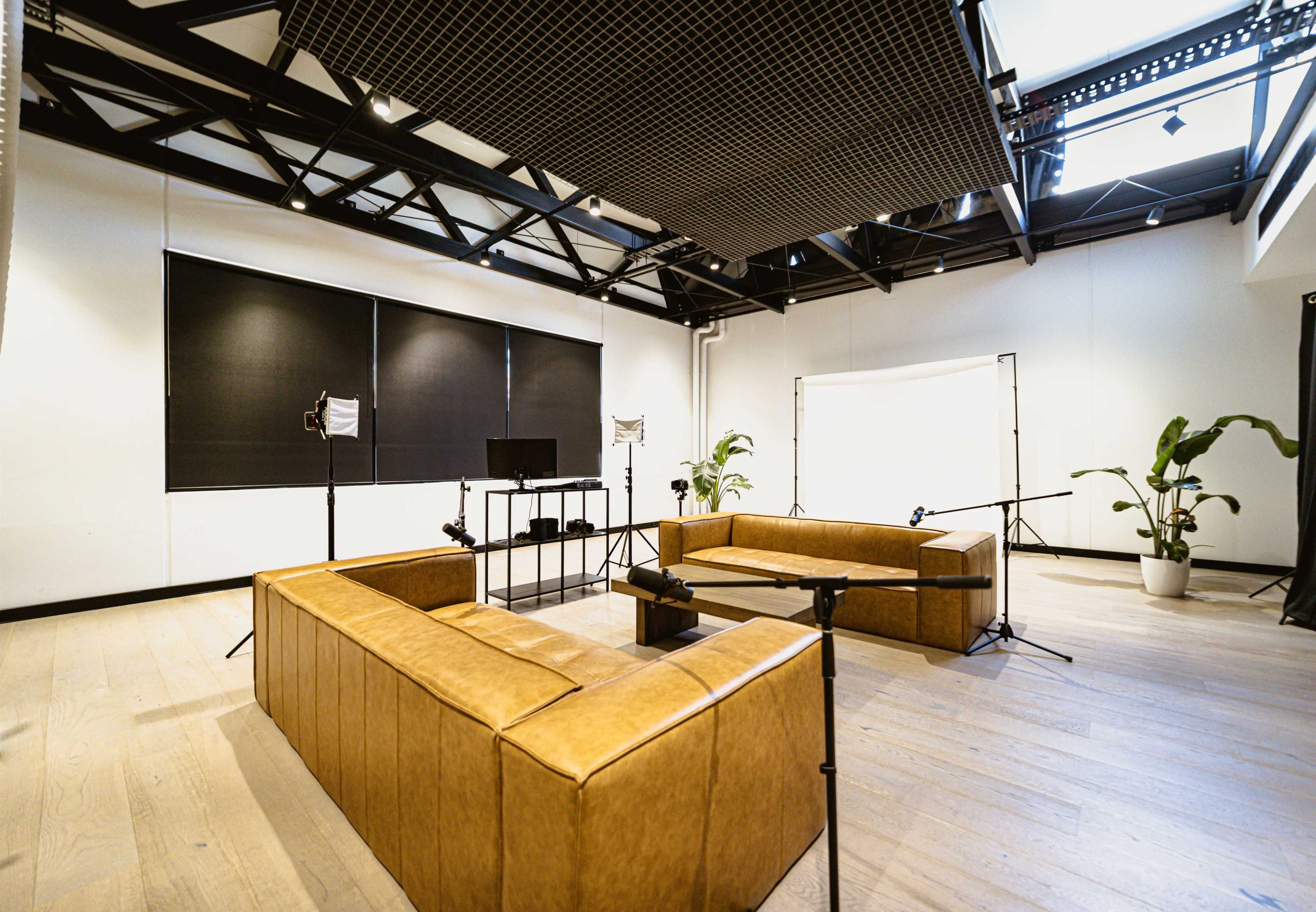 A spacious, well-lit studio equipped with two brown sofas, a coffee table, and multiple lighting setup stands.