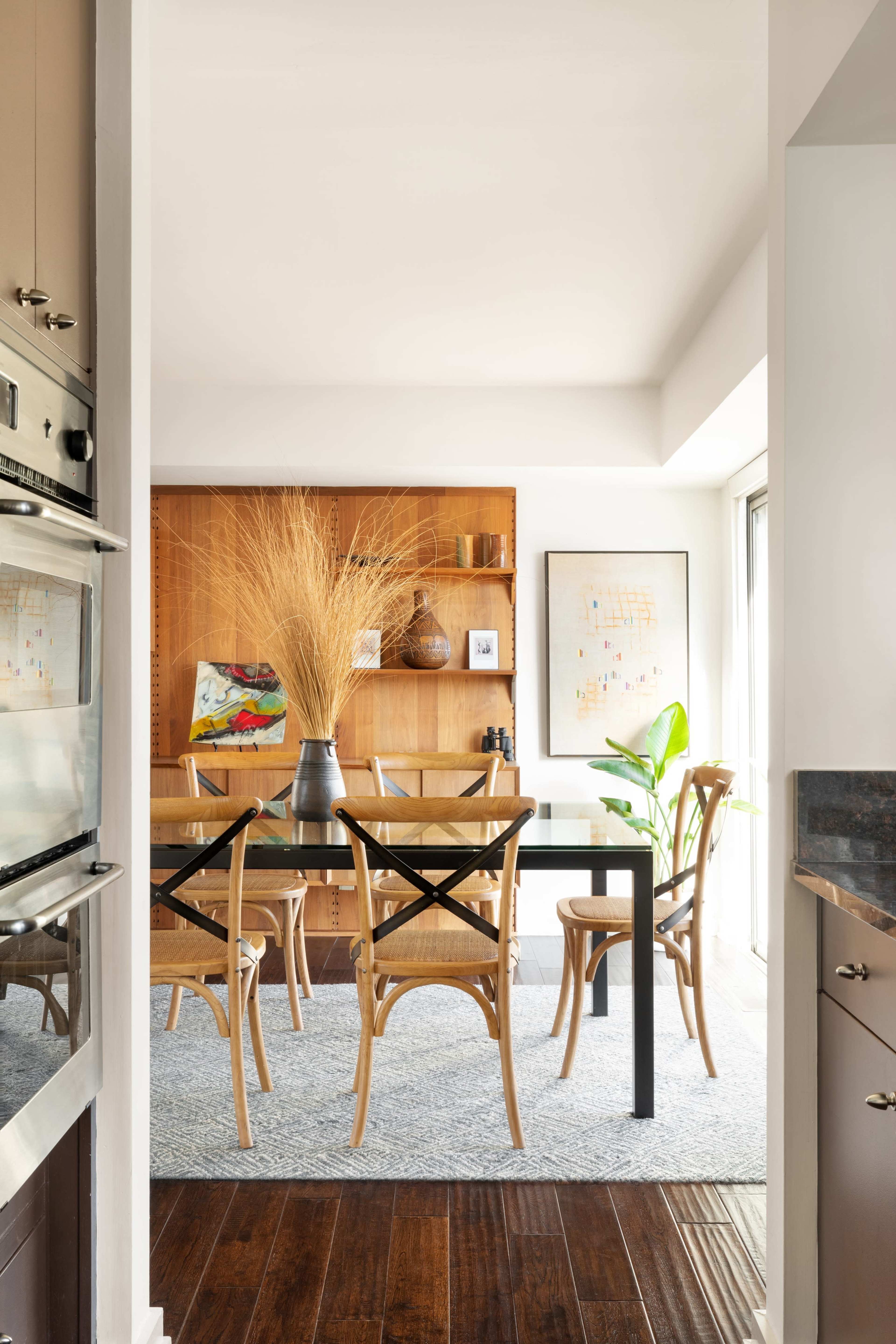 A dining room features a wooden table surrounded by chairs, with a decorative shelf and large vase in the background.