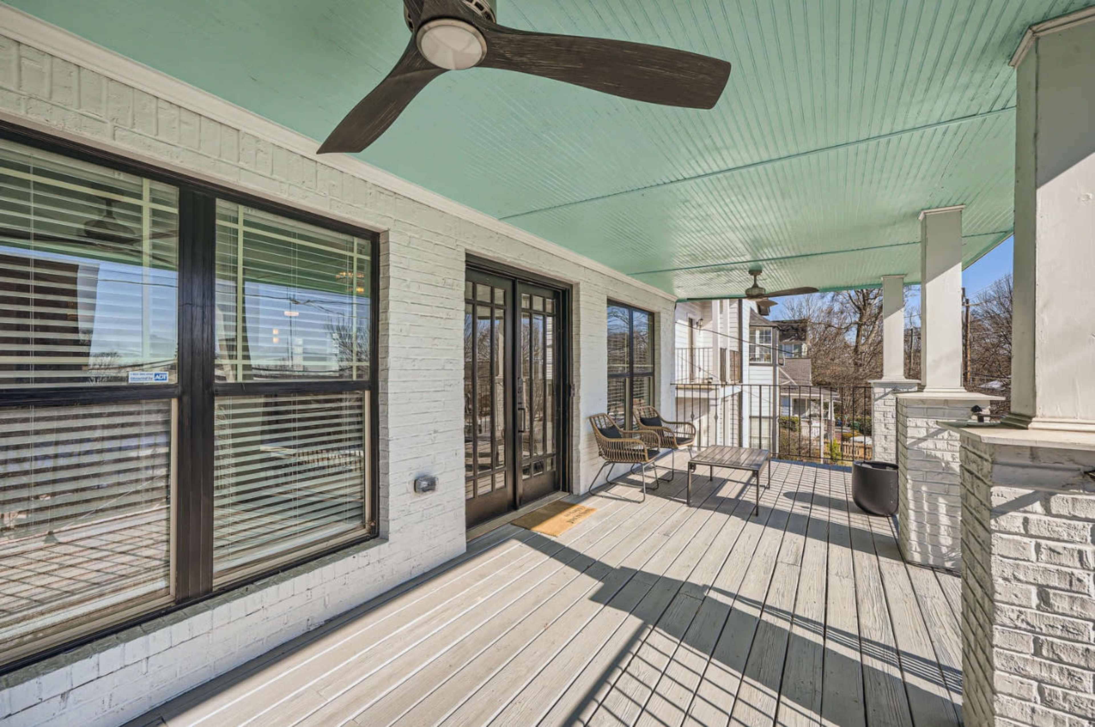 The image shows a covered porch with a light blue ceiling, black sliding doors, and a couple of chairs arranged on the wooden decking.