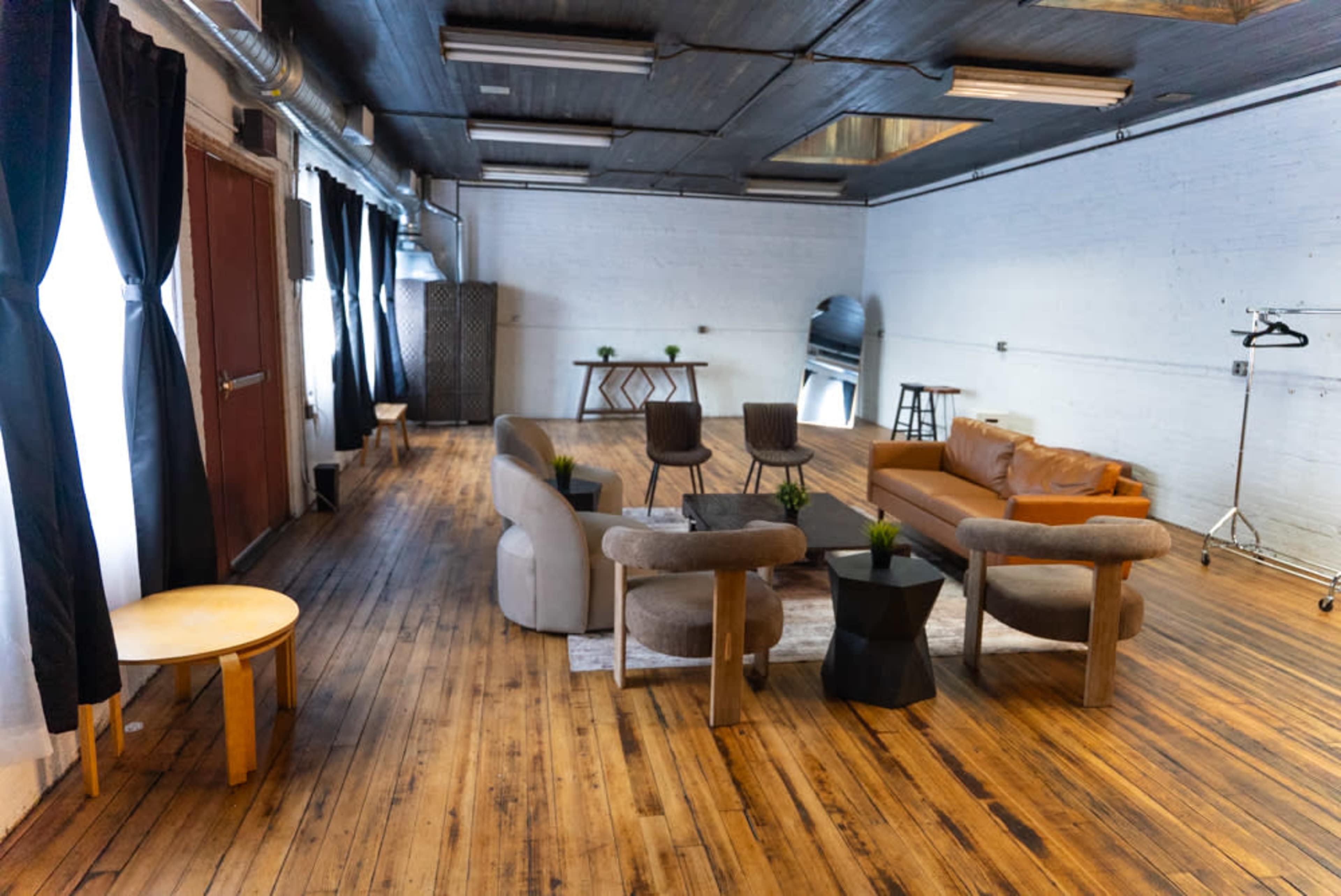 The scene features a spacious, well-lit room with wooden flooring, furnished with a mix of seating and tables, including a leather couch and several chairs around a small black table.