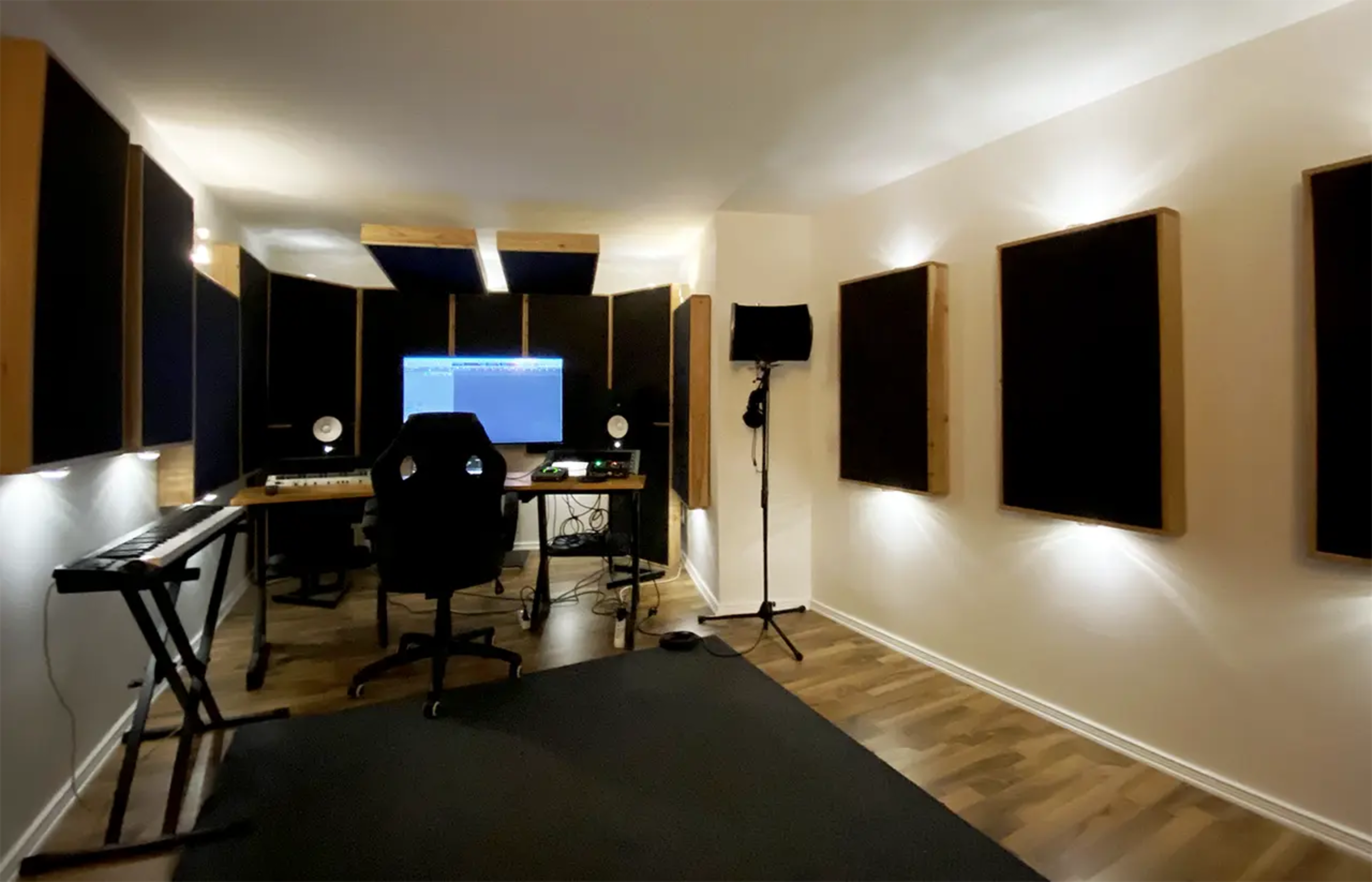 The image shows a music recording studio with soundproofing panels on the walls, a desk with a computer, a keyboard, and a microphone stand.