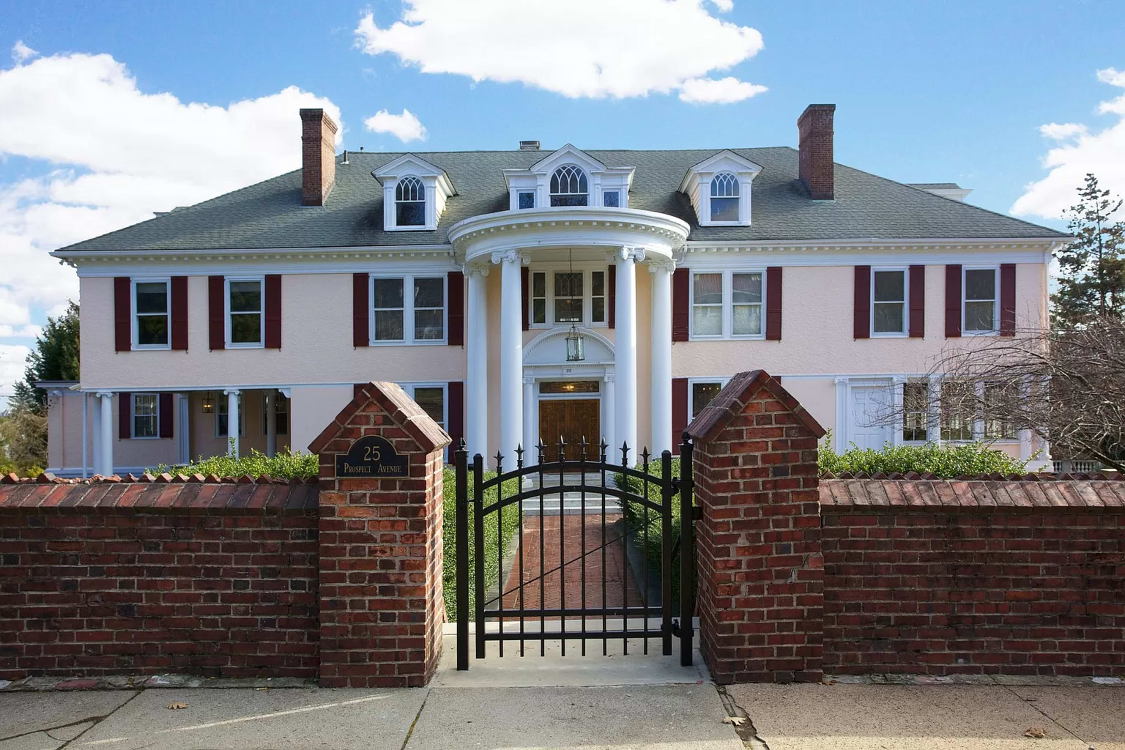 A large, pastel-colored mansion with white columns, red shutters, and a wrought iron gate, surrounded by a brick wall and a neatly landscaped yard.