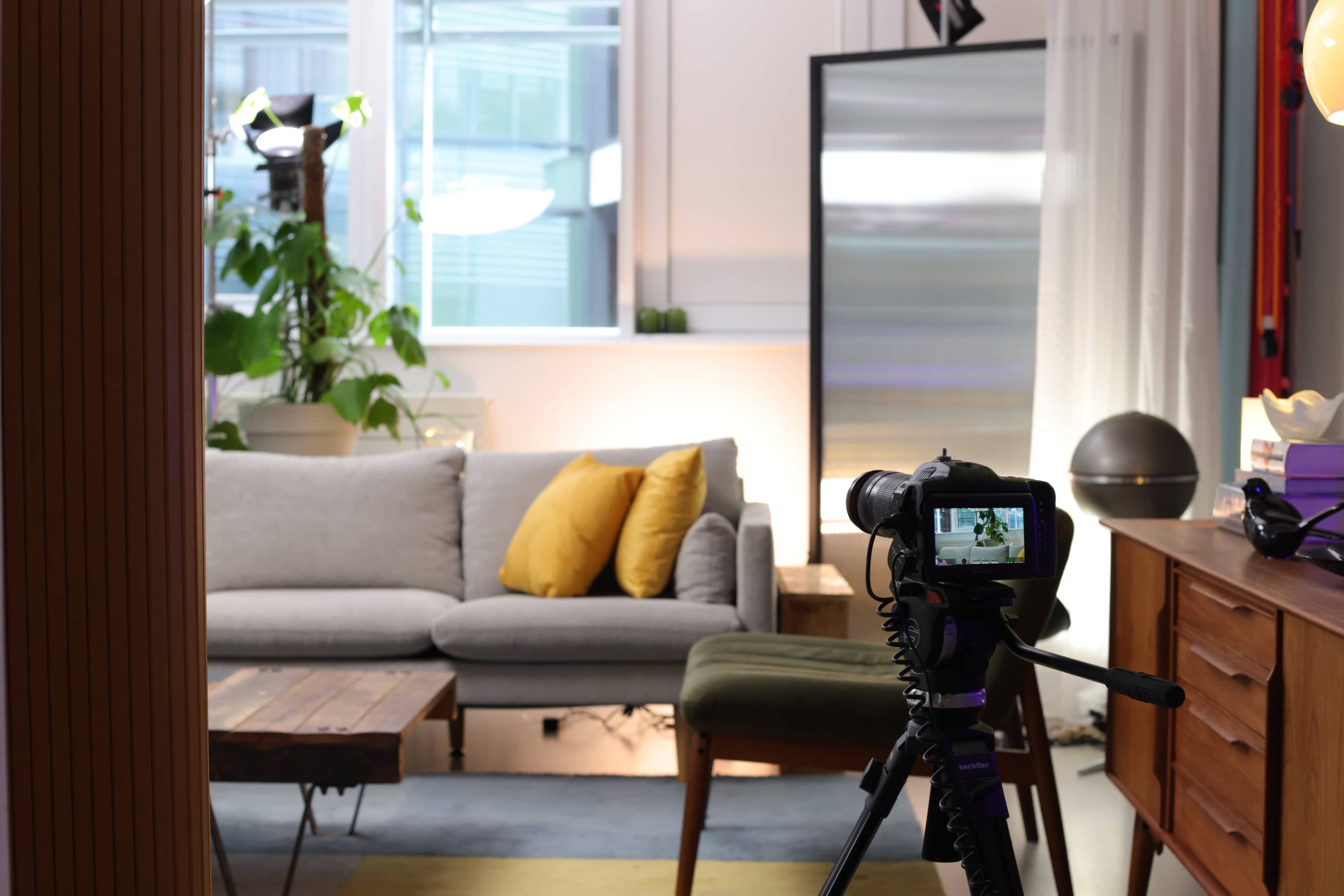 A camera on a tripod is positioned in front of a cozy living room setup featuring a gray sofa with yellow pillows, a wooden coffee table, and a large window with greenery.