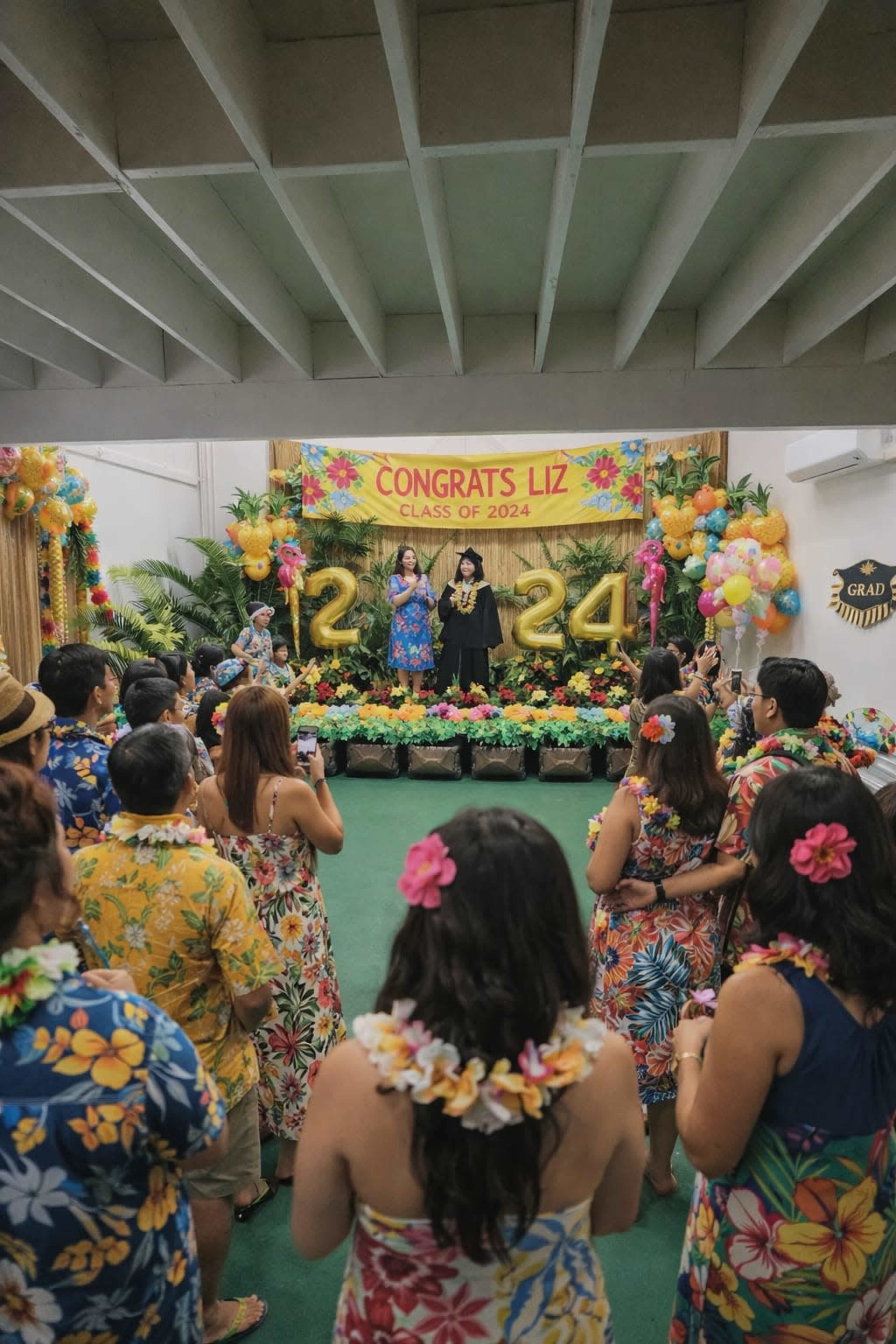 A group of people in colorful floral shirts gathers in a decorated venue to celebrate a graduation, with someone speaking on stage.