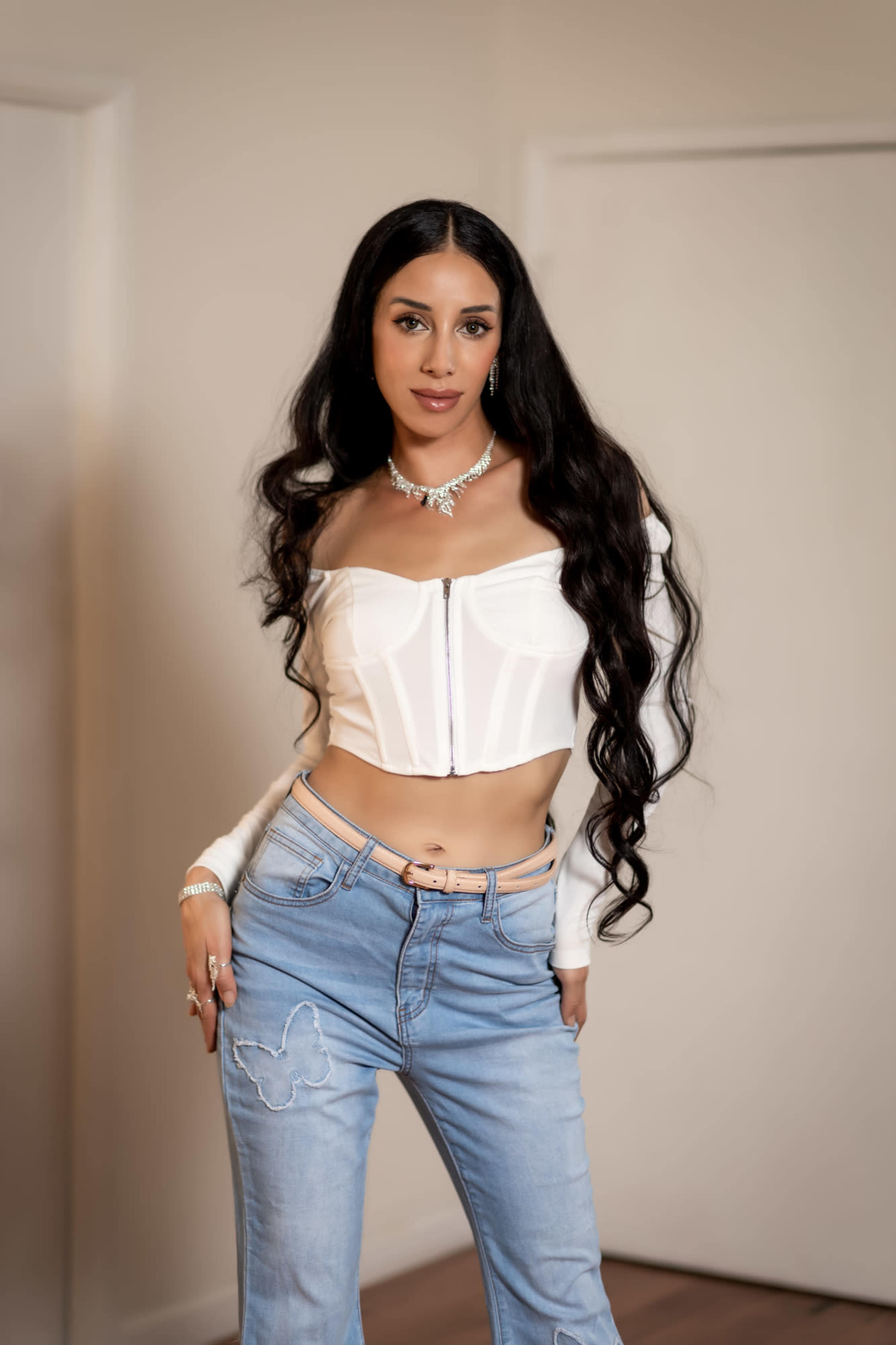 A woman with long, wavy hair poses in a fitted white crop top and blue jeans against a plain background.