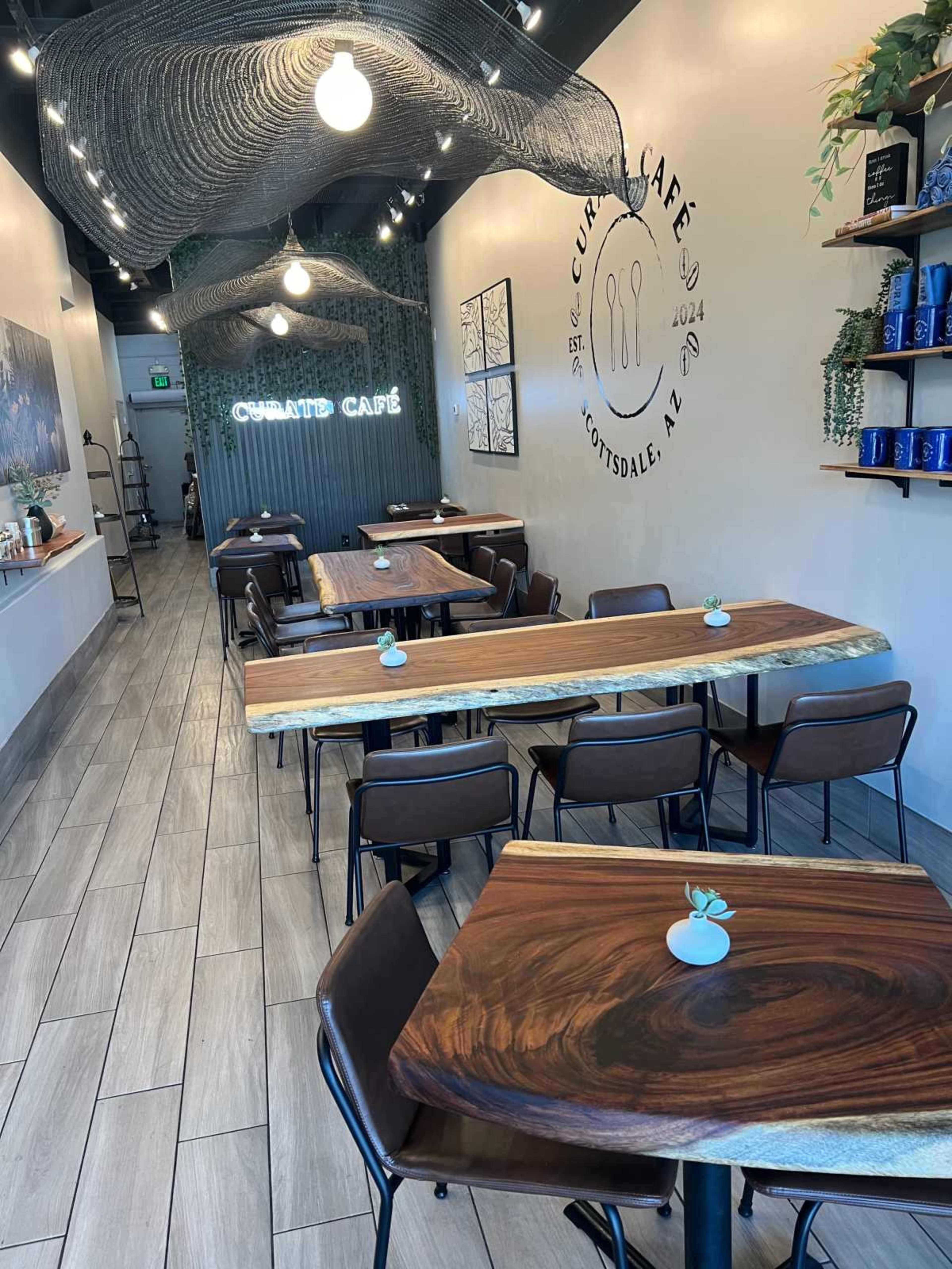 A modern café interior with wooden tables, brown chairs, and a minimalist design featuring a decorative wall with the café's name and plants.