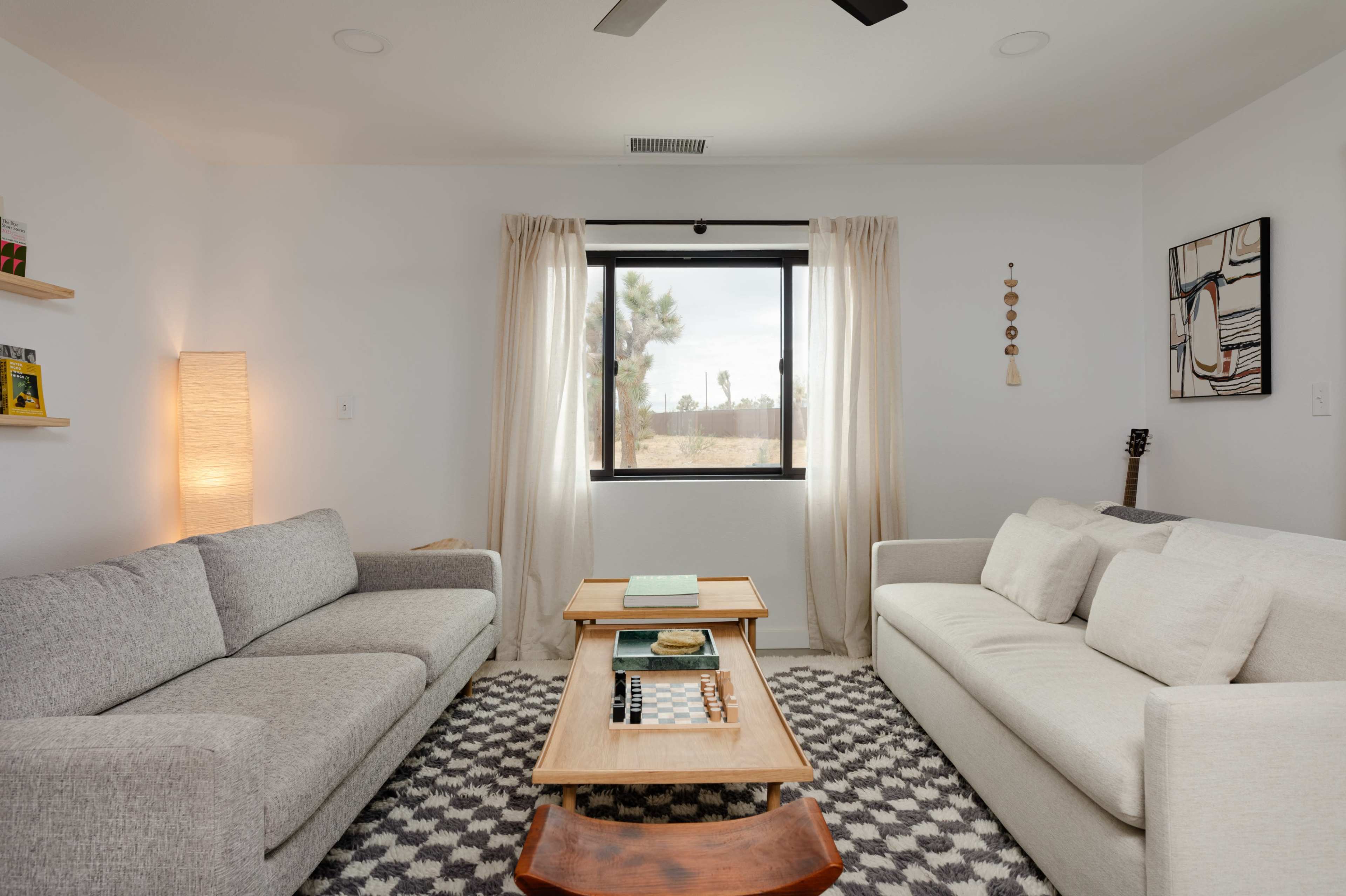The living room features two sofas facing each other, a wooden coffee table between them, and a large window with sheer curtains offering a view of the outdoors.