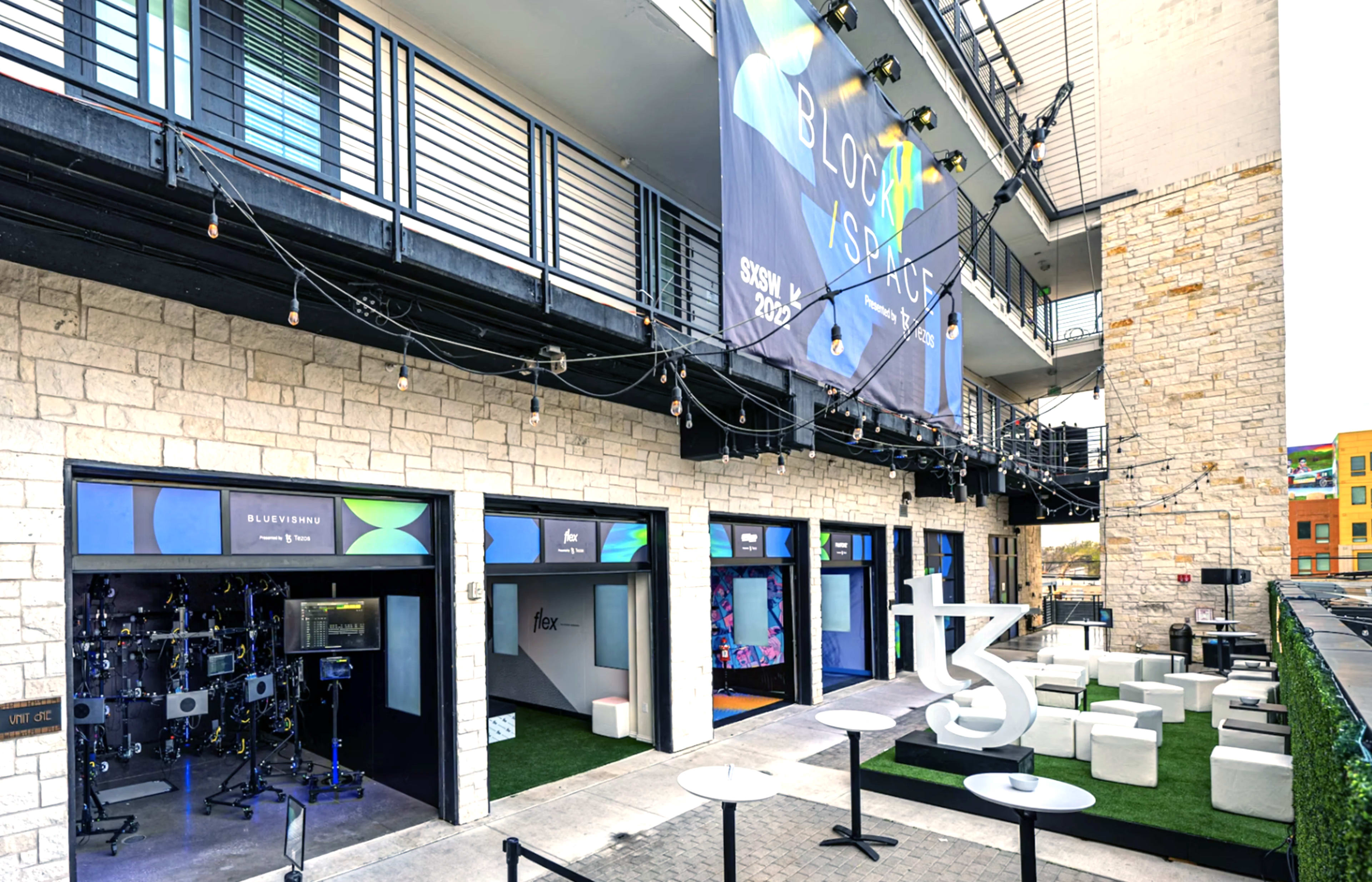 The image shows a modern outdoor venue with a stone building, featuring several booths and a large banner, with seating areas visible.