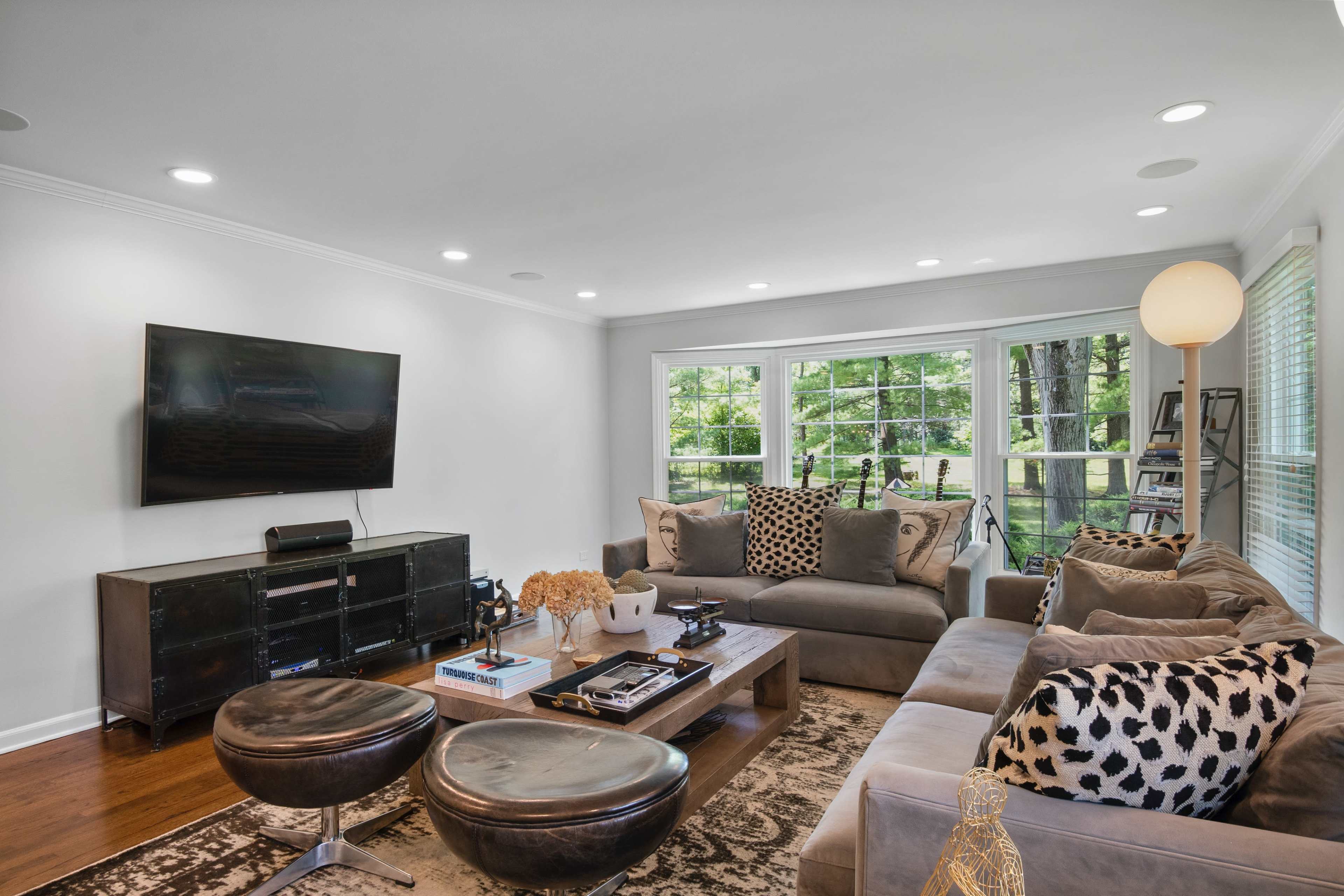 The living room features a large sectional sofa, a coffee table, and a TV mounted on the wall, with large windows providing a view of the outside.