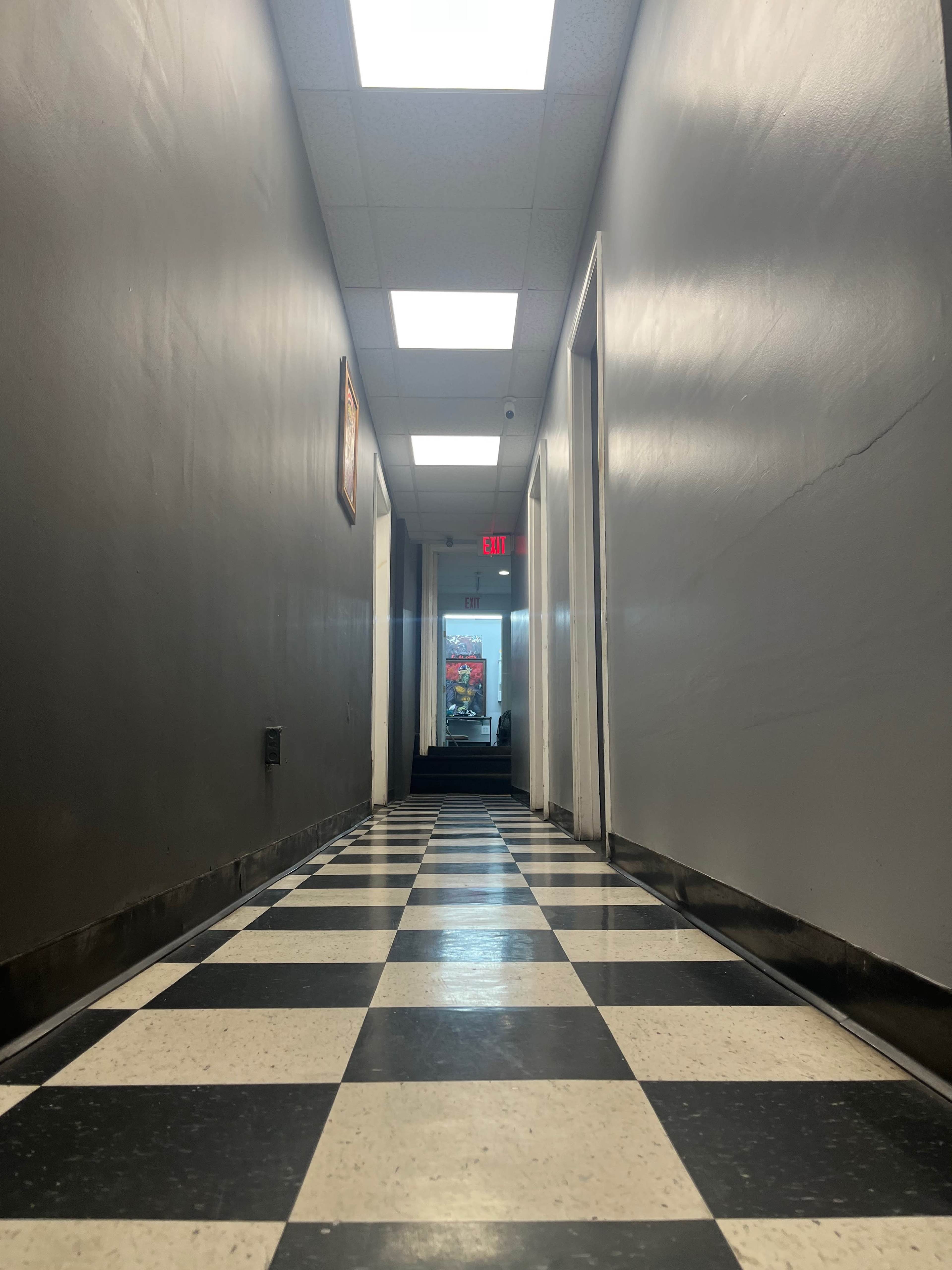 A long corridor with black and white checkered flooring leads to a glass door at the far end, illuminated by ceiling lights.