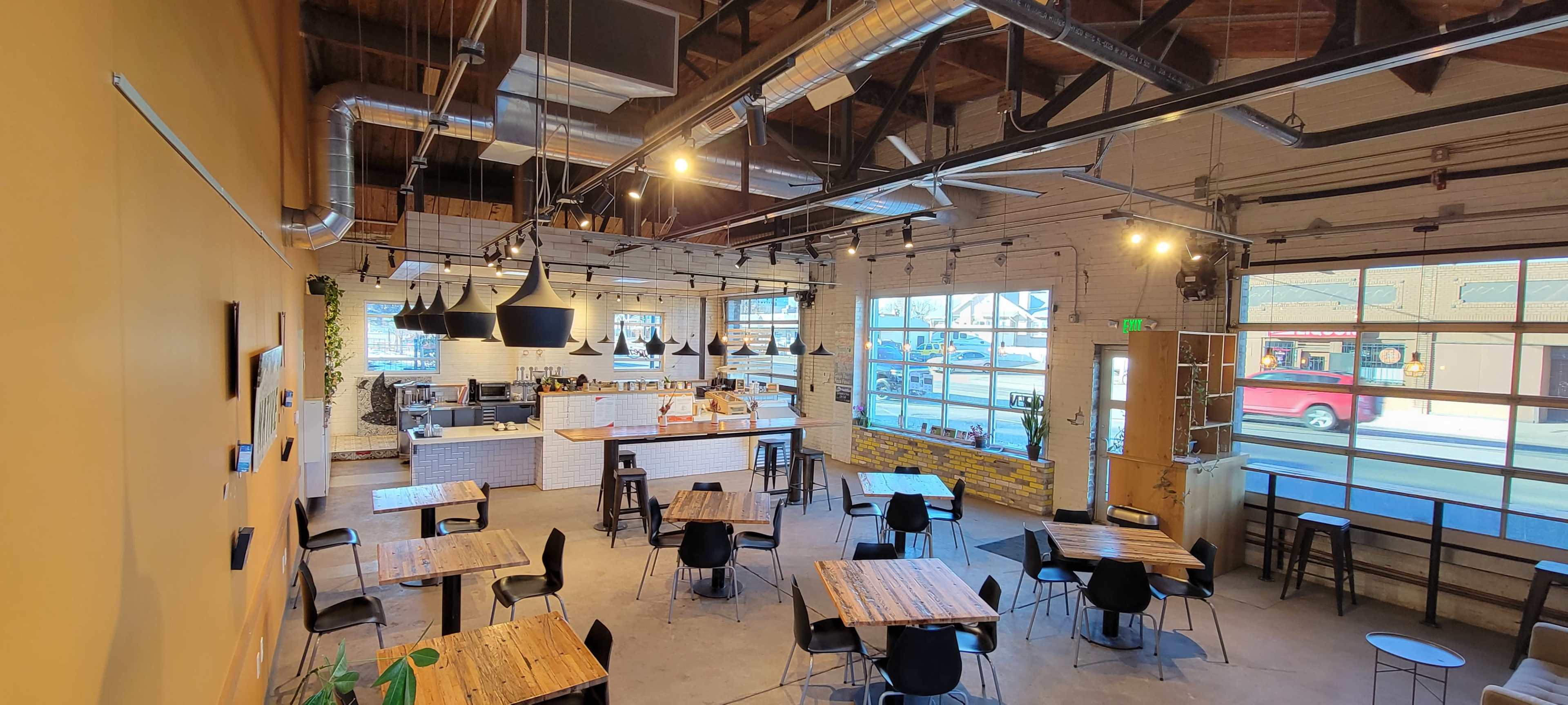 An interior view of a modern café featuring wooden tables and black chairs, with a service counter in the background and large windows allowing natural light to enter.