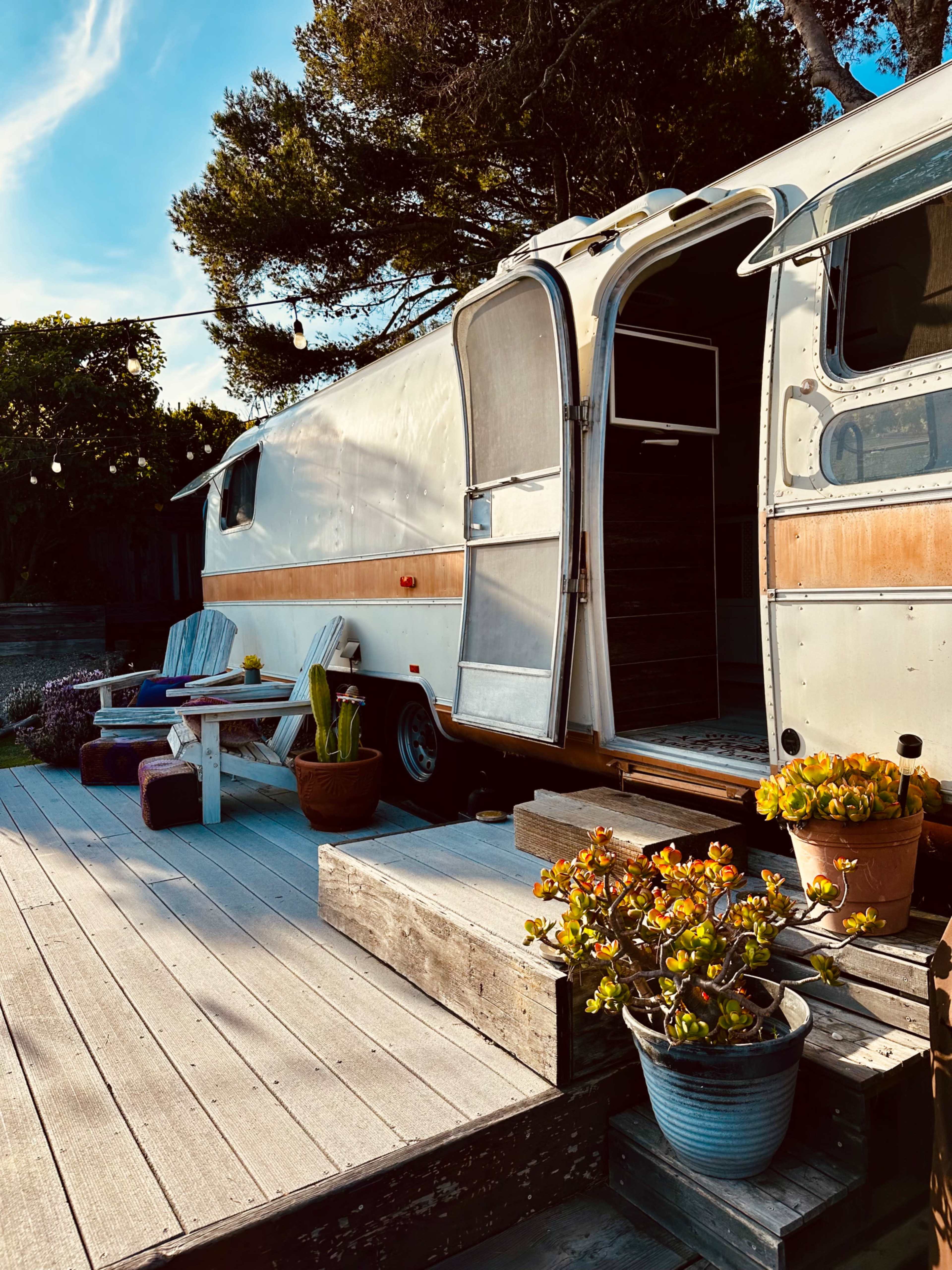 An Airstream trailer is parked next to a wooden deck adorned with potted plants and outdoor seating.