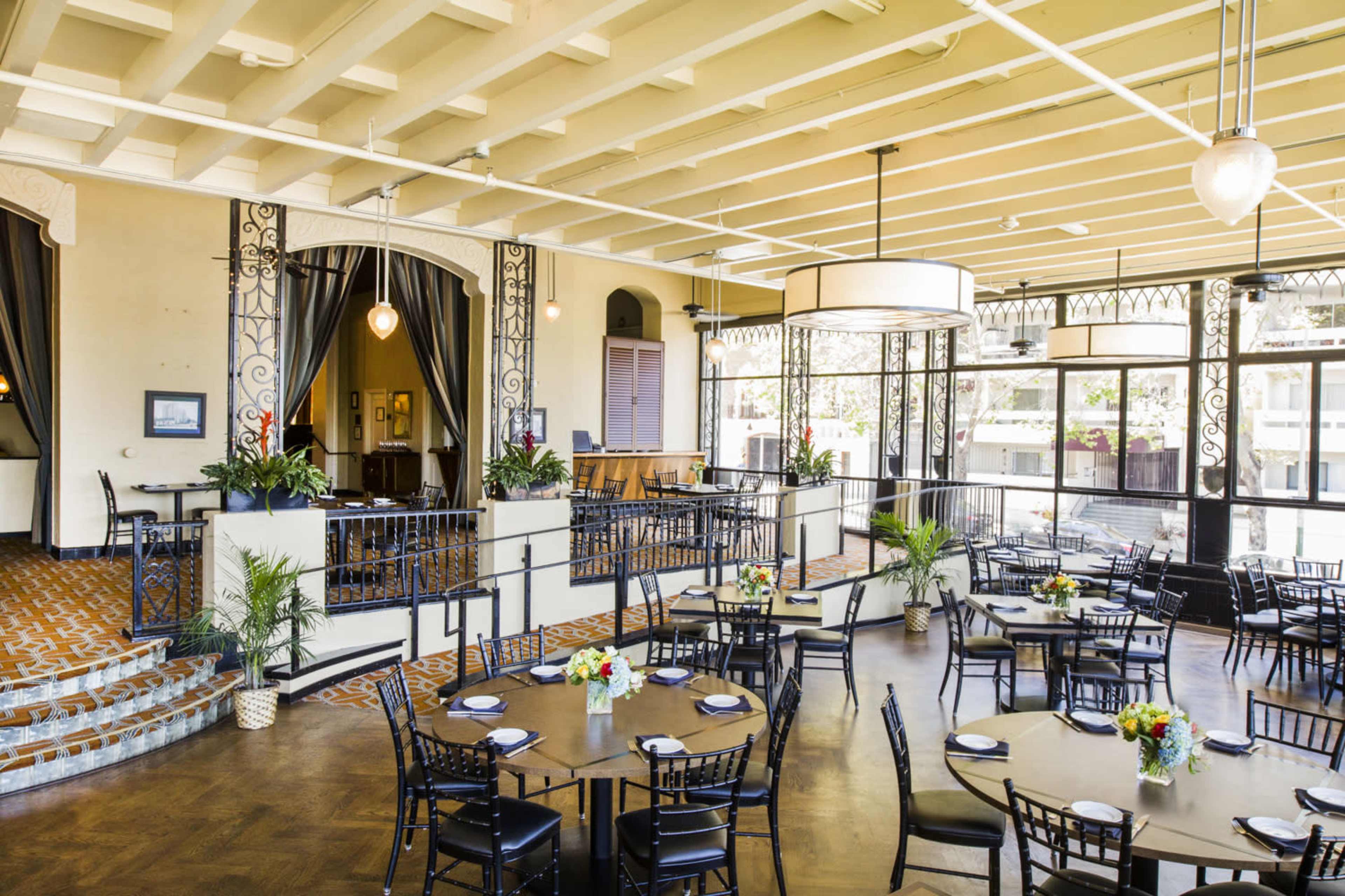 The image shows a spacious restaurant interior with round tables set for dining, surrounded by decorative plants and elegant light fixtures.