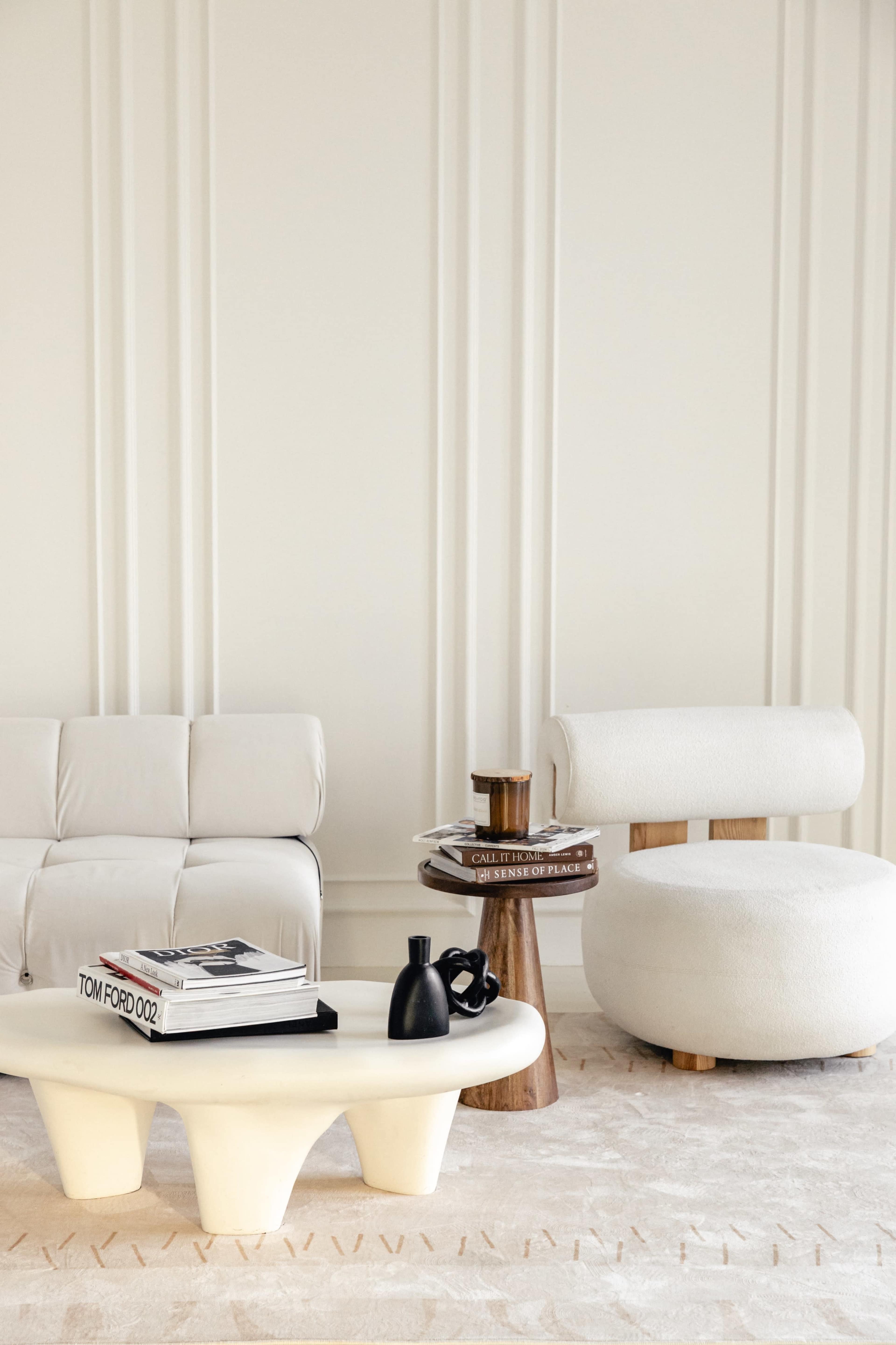The image features a minimalist living room setting with a light-colored sofa, a rounded armchair, a sculptural coffee table, and a side table stacked with books and a small decorative item.