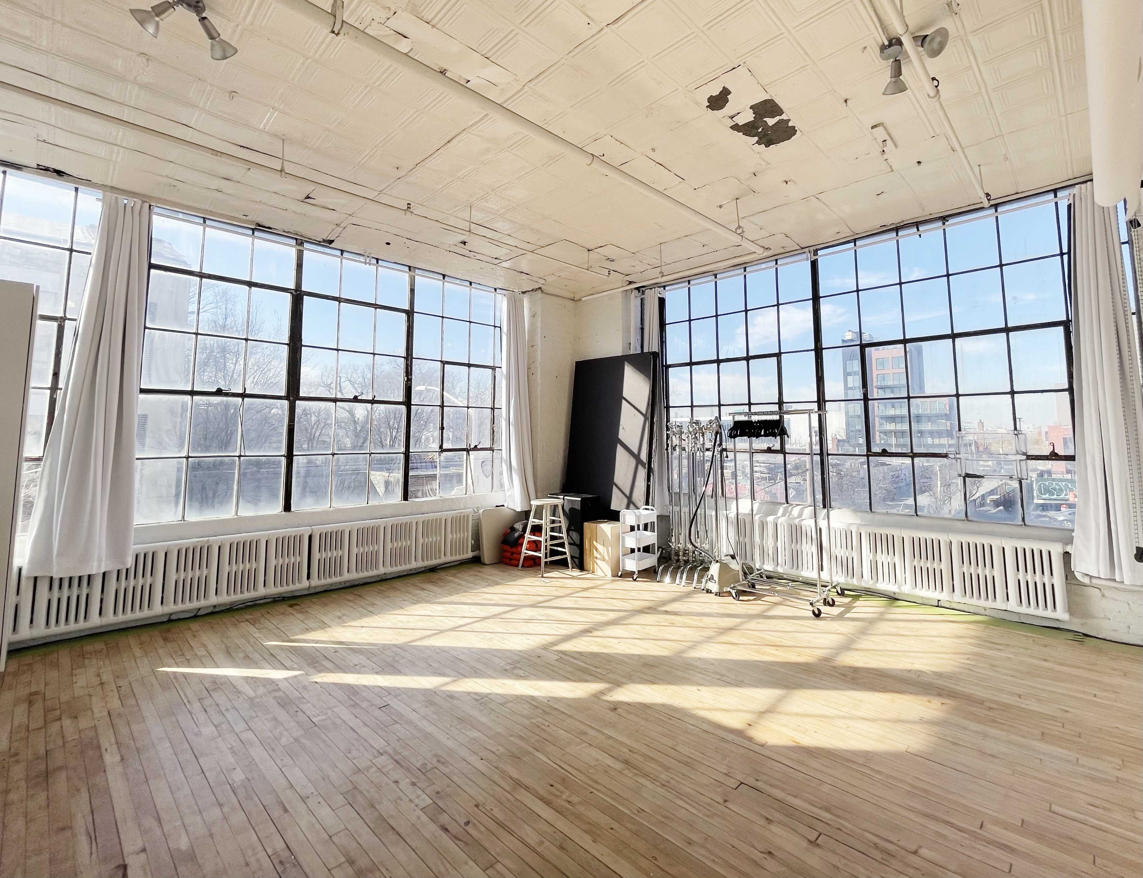 A spacious, sunlit room with large windows, wooden flooring, and minimal furnishings.