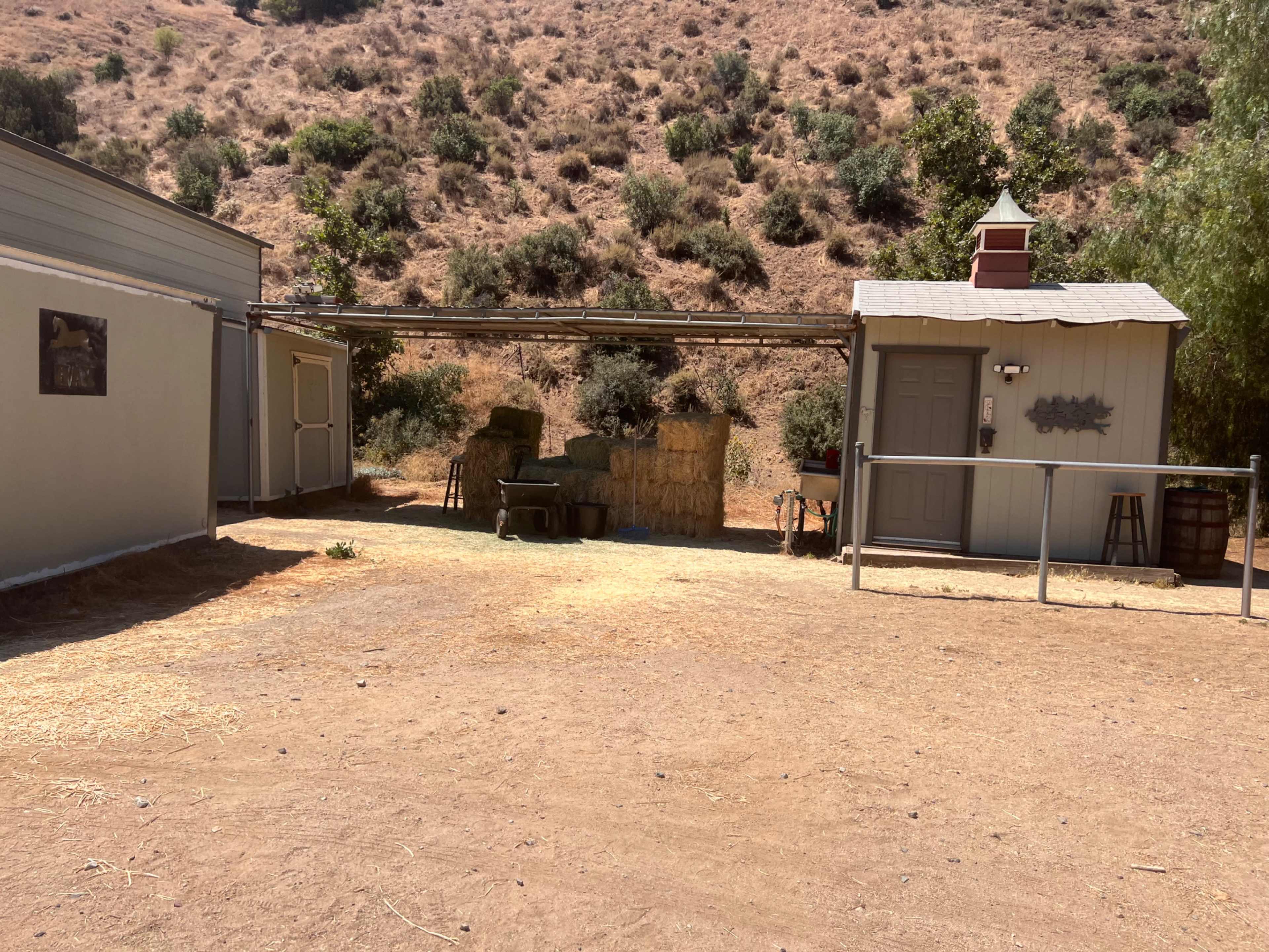 A rustic outdoor area with a small building, hay bales stacked nearby, and a dry, brown landscape in the background.