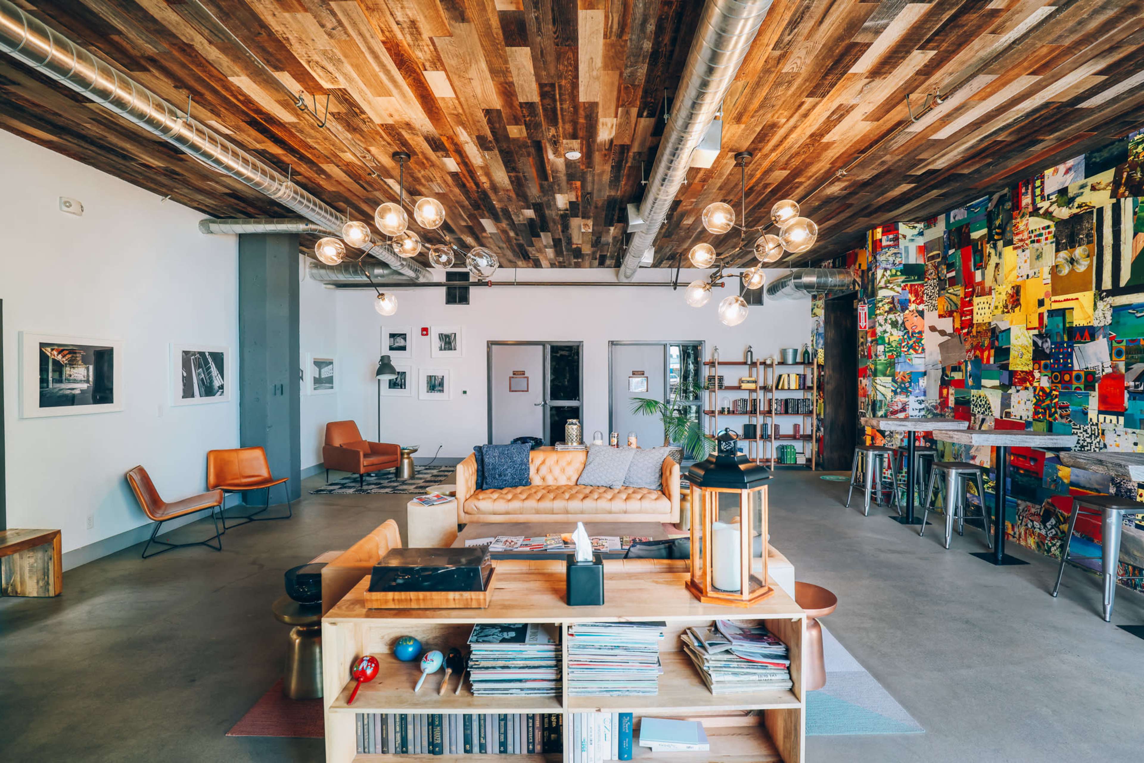 The image shows a modern living space with wooden ceilings, a cozy sofa, bookshelves, and various artworks displayed on the walls.