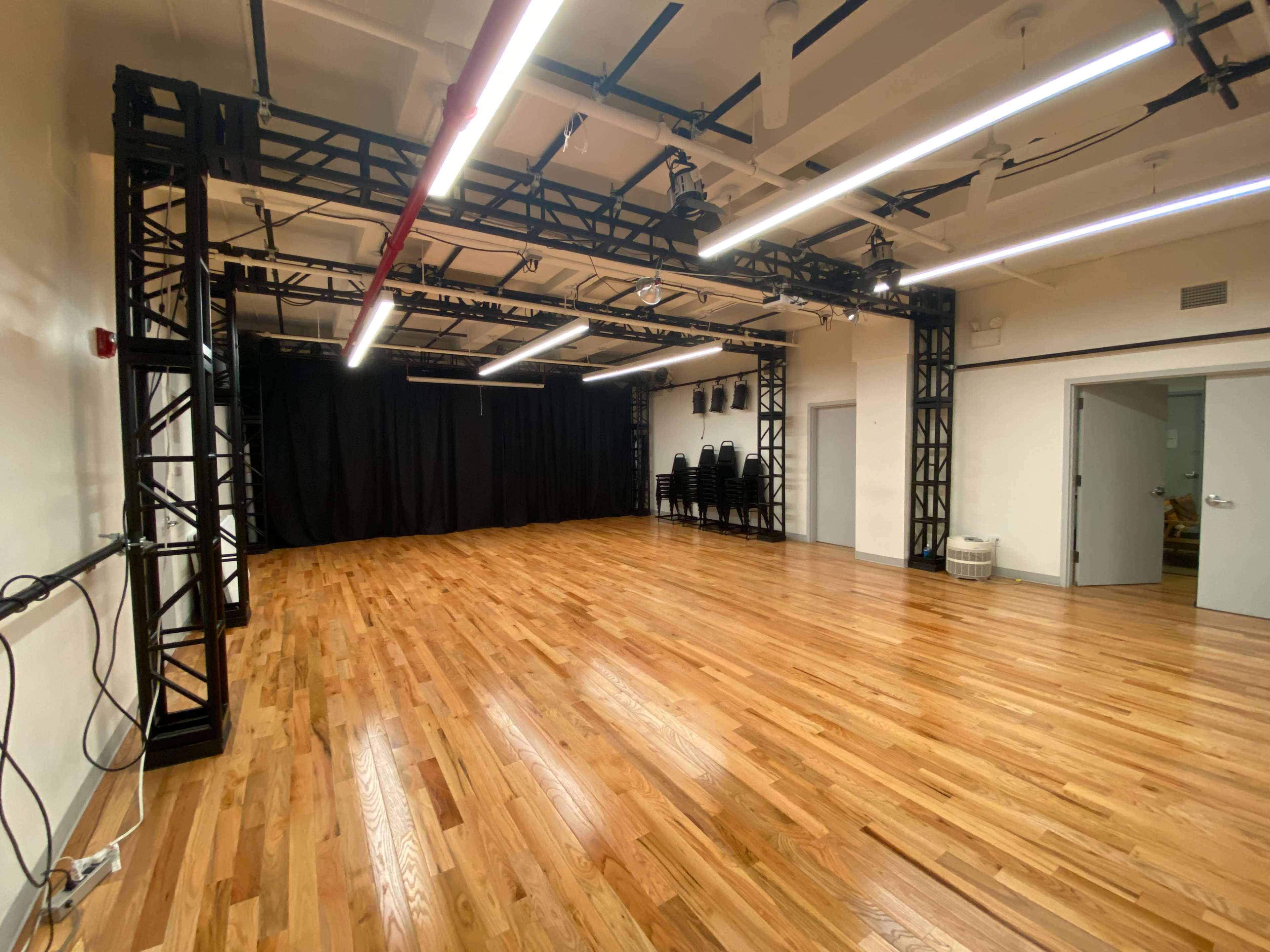 A spacious dance studio with wooden flooring, black staging equipment, and stacked chairs against the wall.